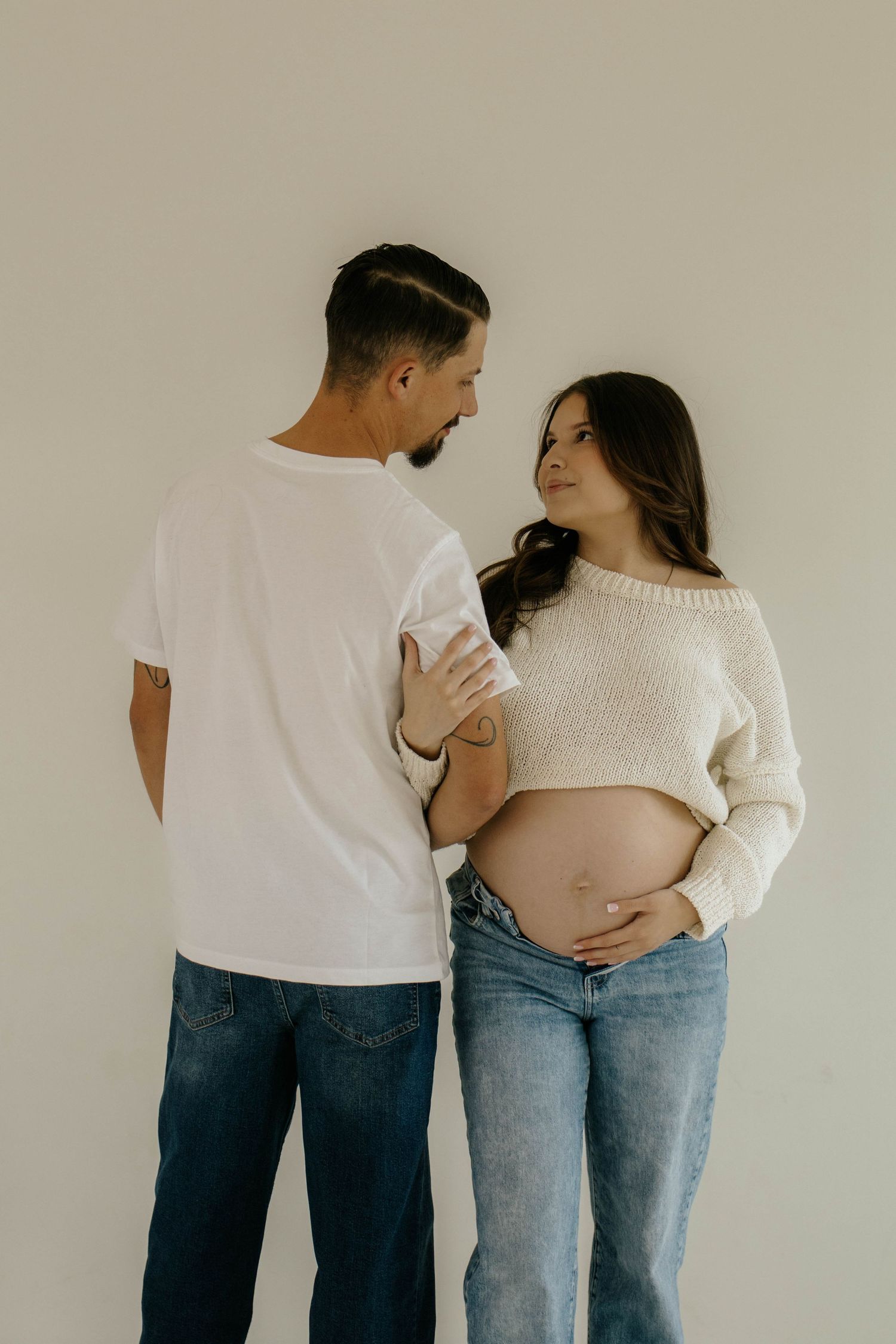 Expecting couple in casual wear sharing an intimate moment while showing off pregnant belly against neutral wall.