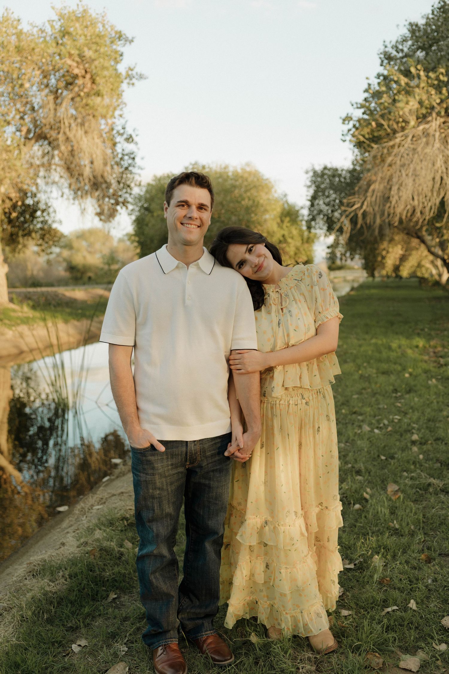 Couple in casual attire poses together on grassy hillside during sunset.