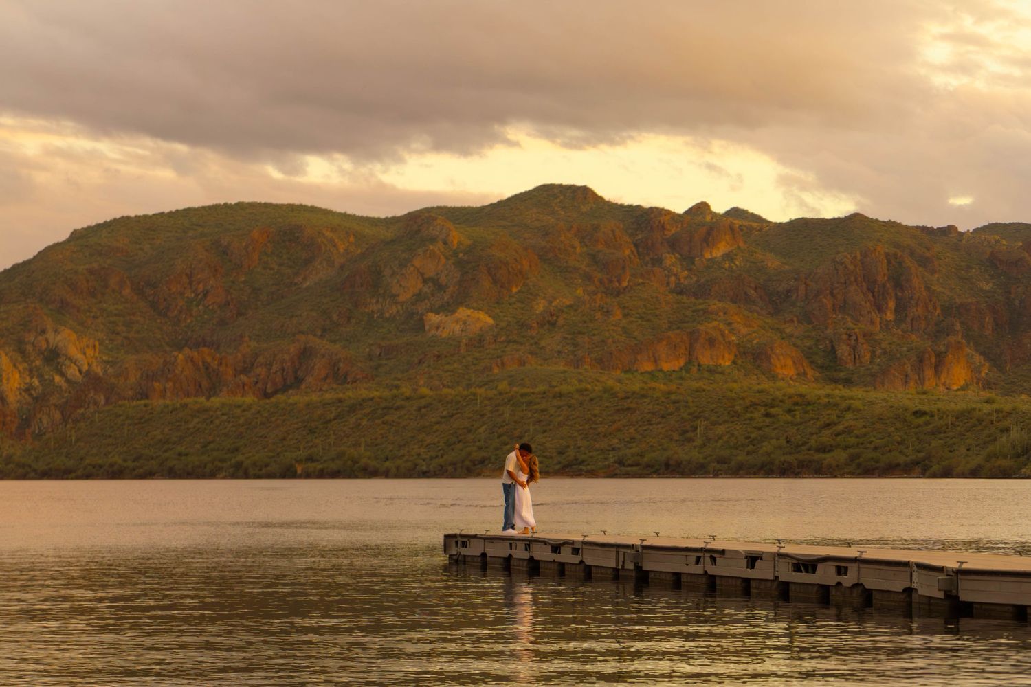 A panoramic view of a coastal landscape with mountains, pier, and lighthouse during golden sunset hour.