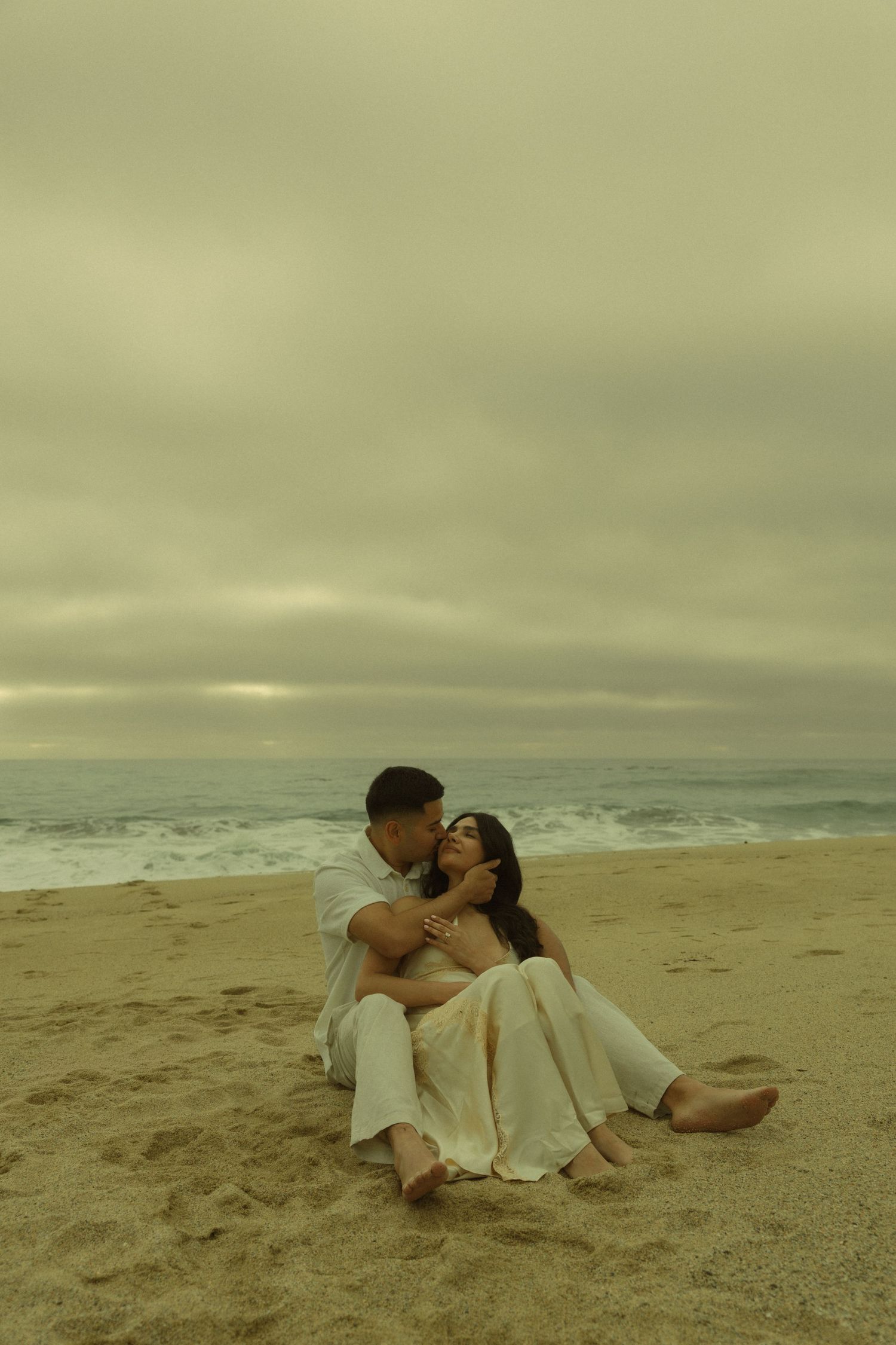 Couple sitting intimately on sandy beach, man kissing woman's head, ocean waves and cloudy sky behind them.