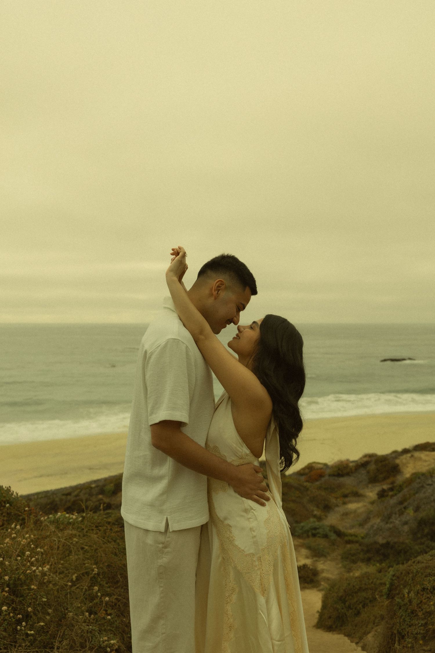 Romantic couple embracing near a coastal cliff with sandy beach and ocean waves in the background on an overcast day.