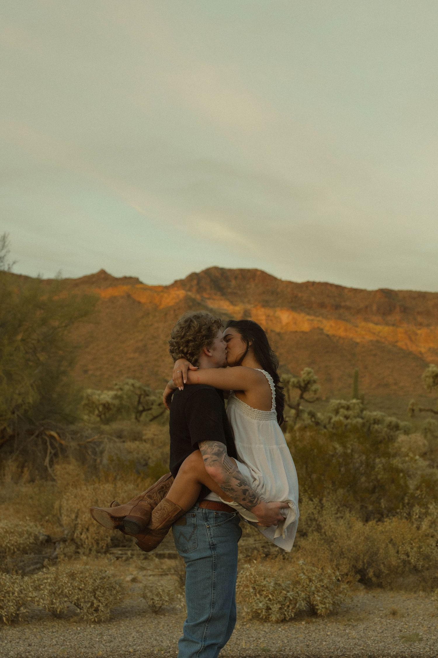 Couple sharing a romantic kiss in the desert at sunset, man lifting woman in white dress with mountains behind.