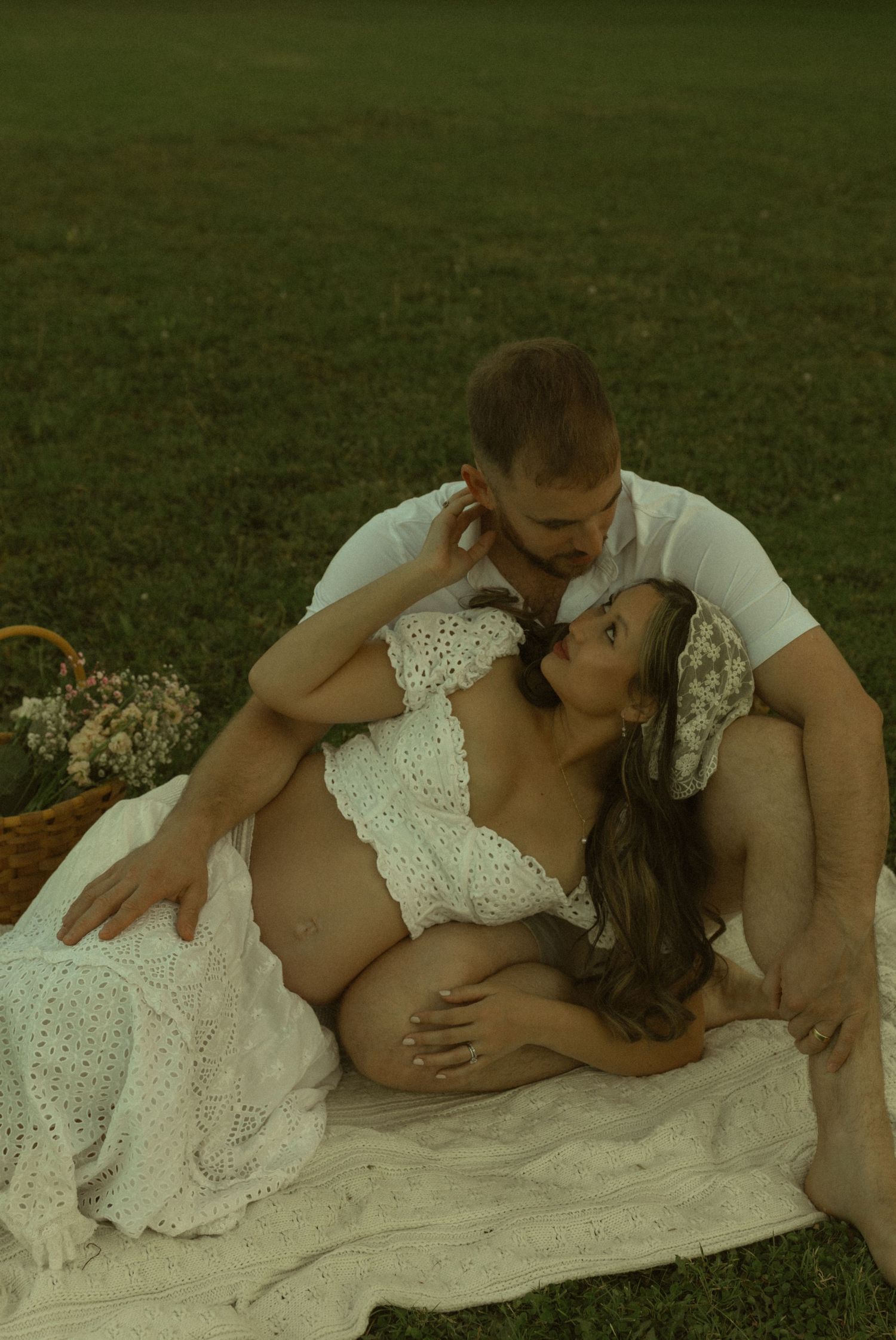Couple sharing a tender moment outdoors on a blanket, pregnant woman in white lace dress cradled by partner on green grass.