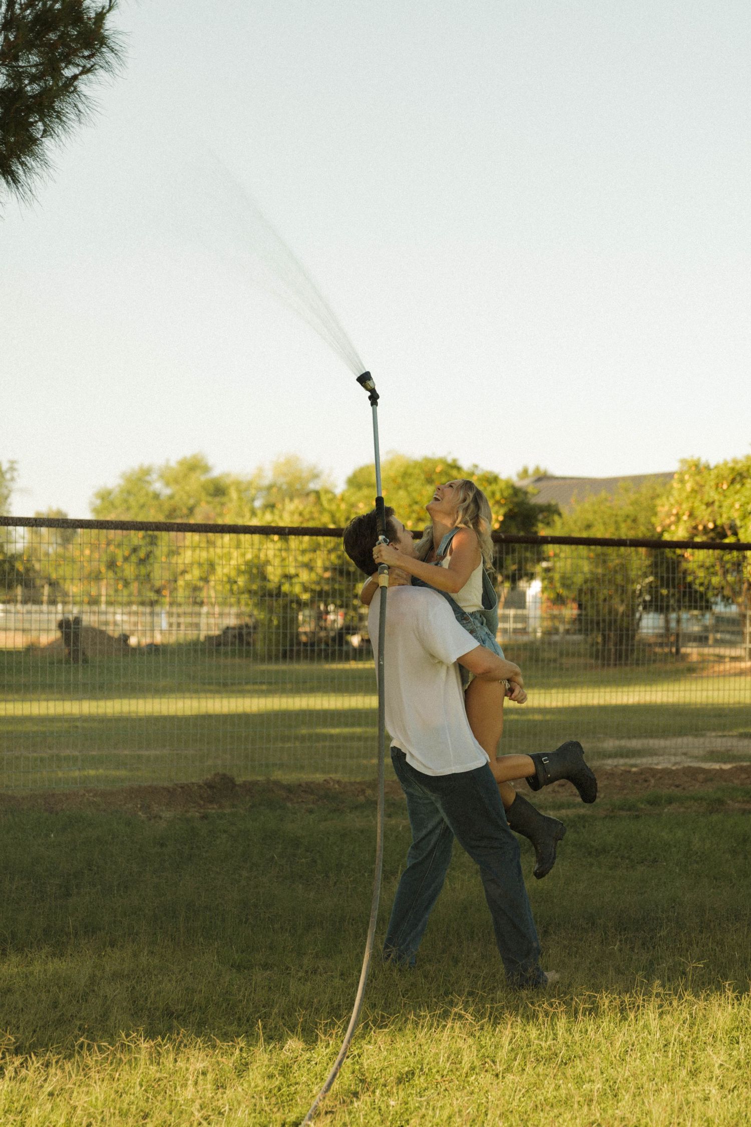 Couple playing with a garden hose at golden hour, man lifting woman as water sprays upward in a sunny backyard.