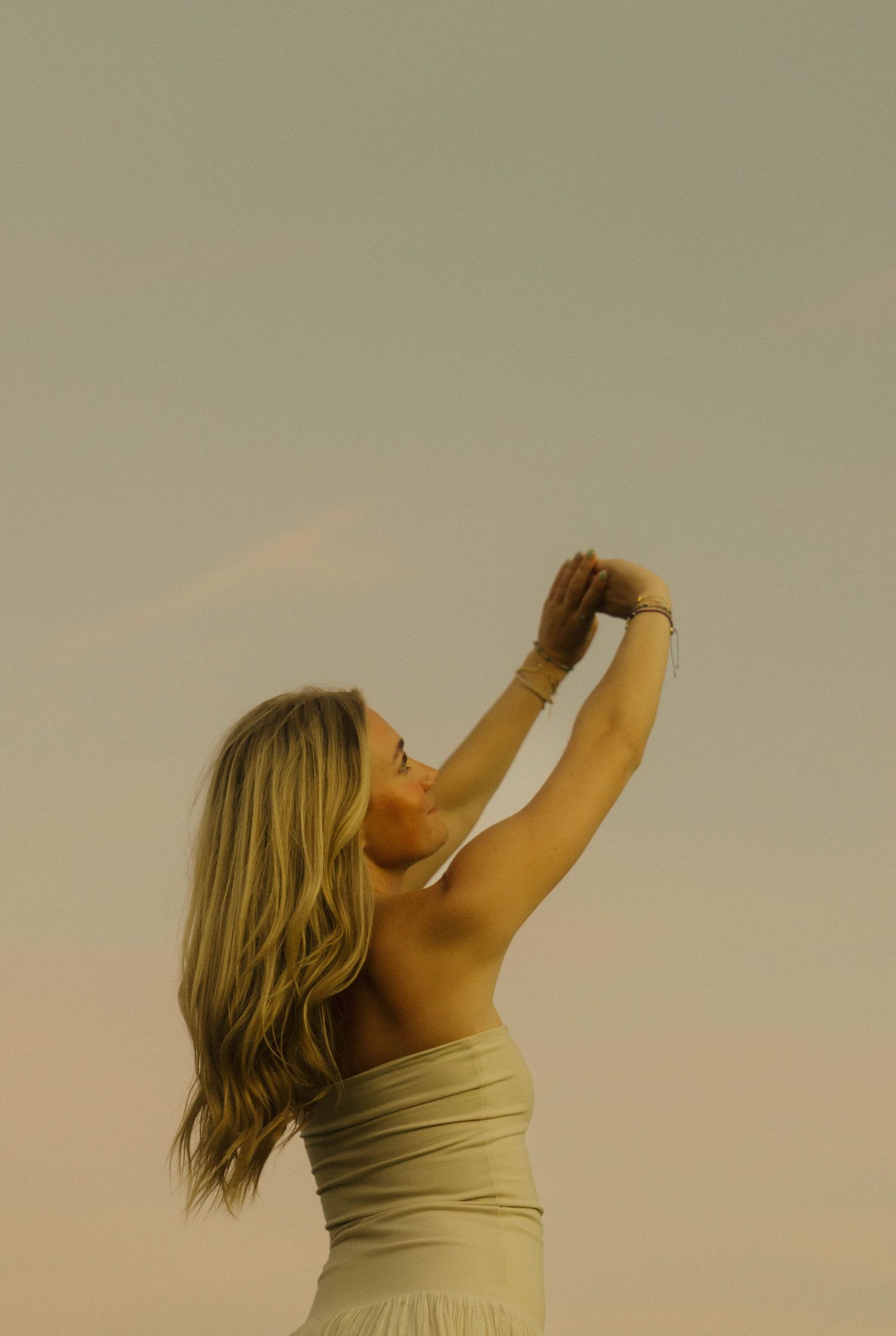 Blonde woman in strapless dress reaching arms overhead against golden sunset sky.