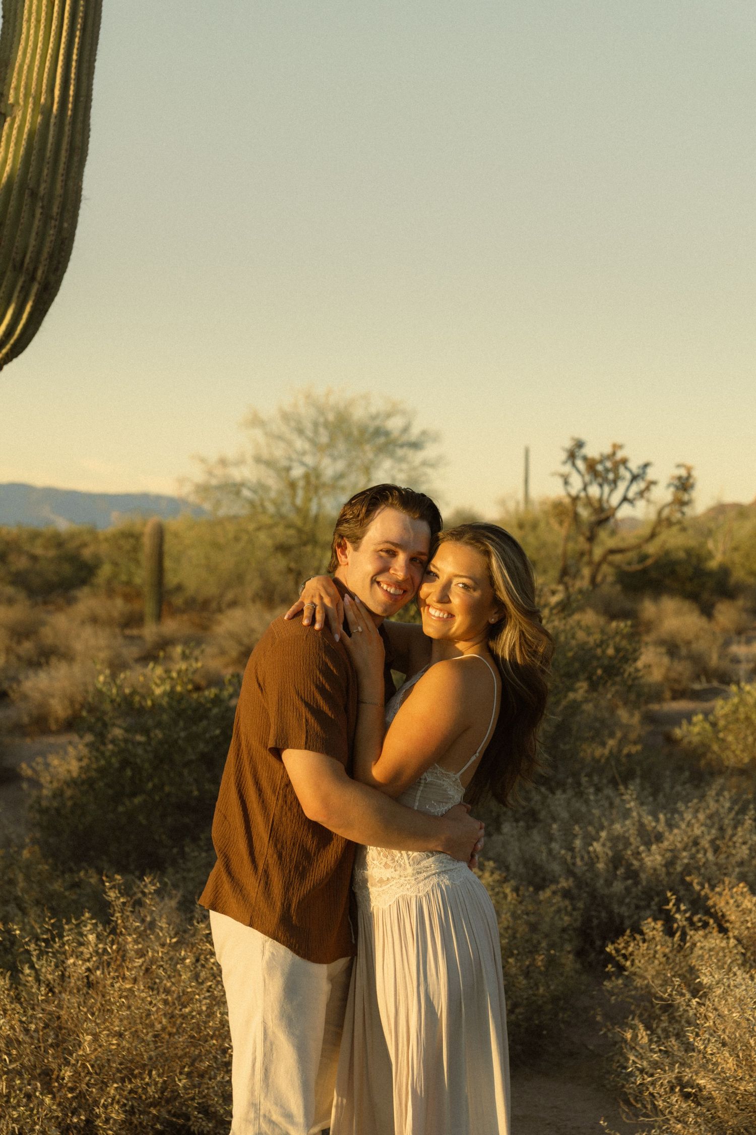 Couple embracing at golden hour in Arizona desert with saguaro cacti and mountains in background.