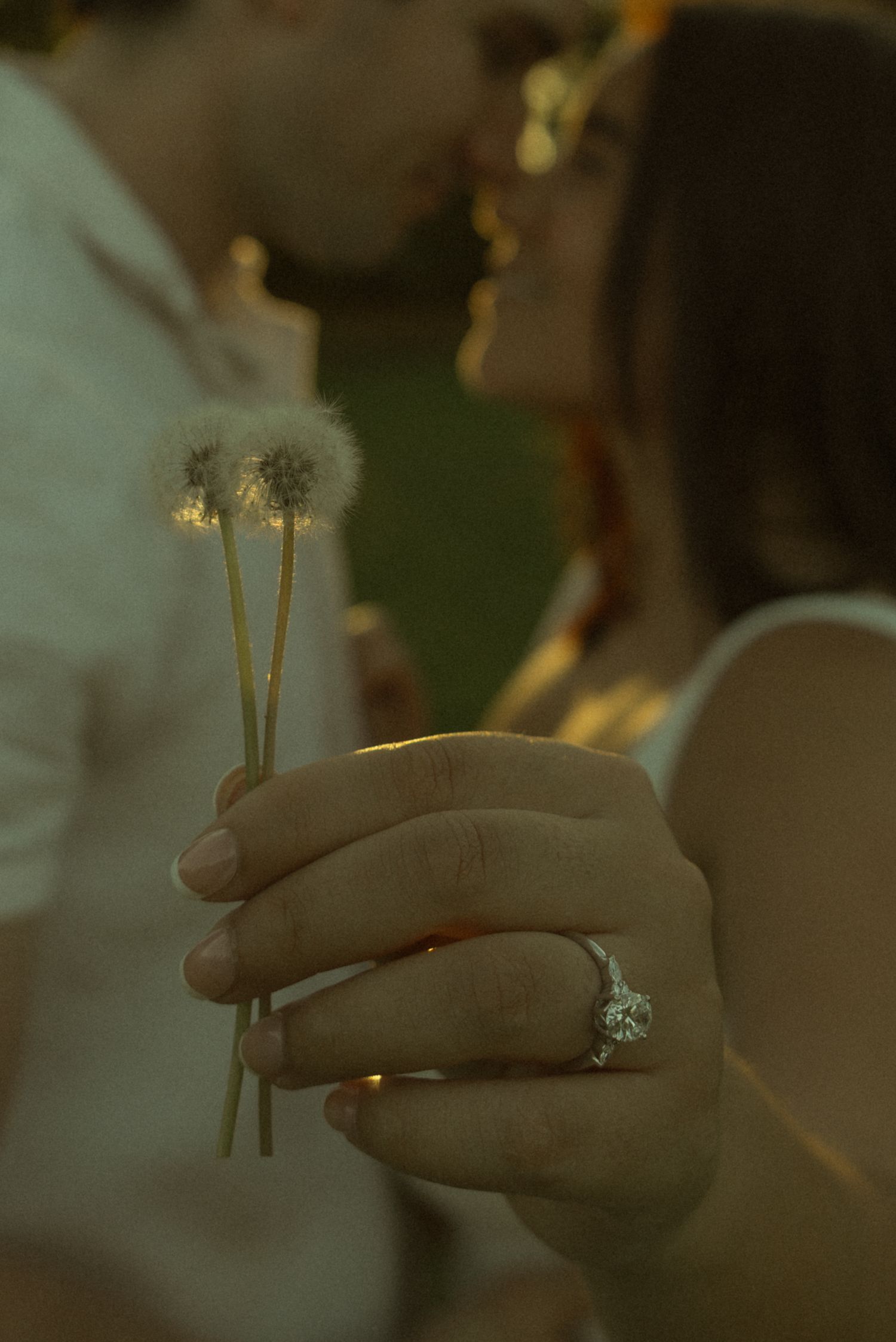 A couple kisses in the background while a hand holding dandelions displays a sparkling diamond engagement ring.
