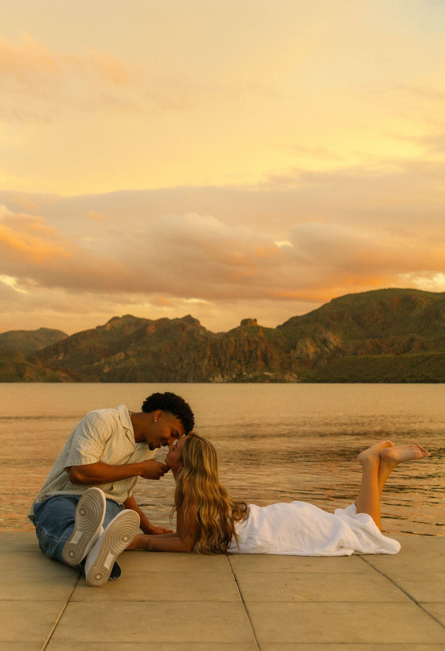 A couple shares a romantic moment at sunset on a stone platform overlooking a serene mountain lake.