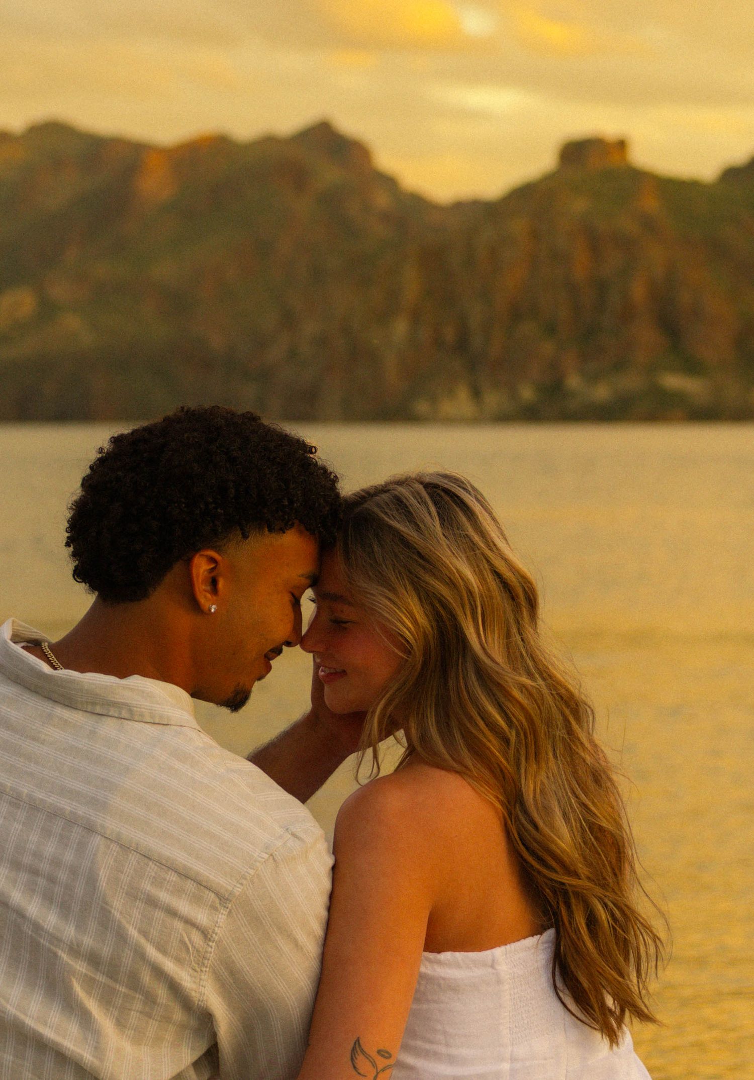 A romantic couple shares an intimate moment at sunset by the water with mountains in the background.