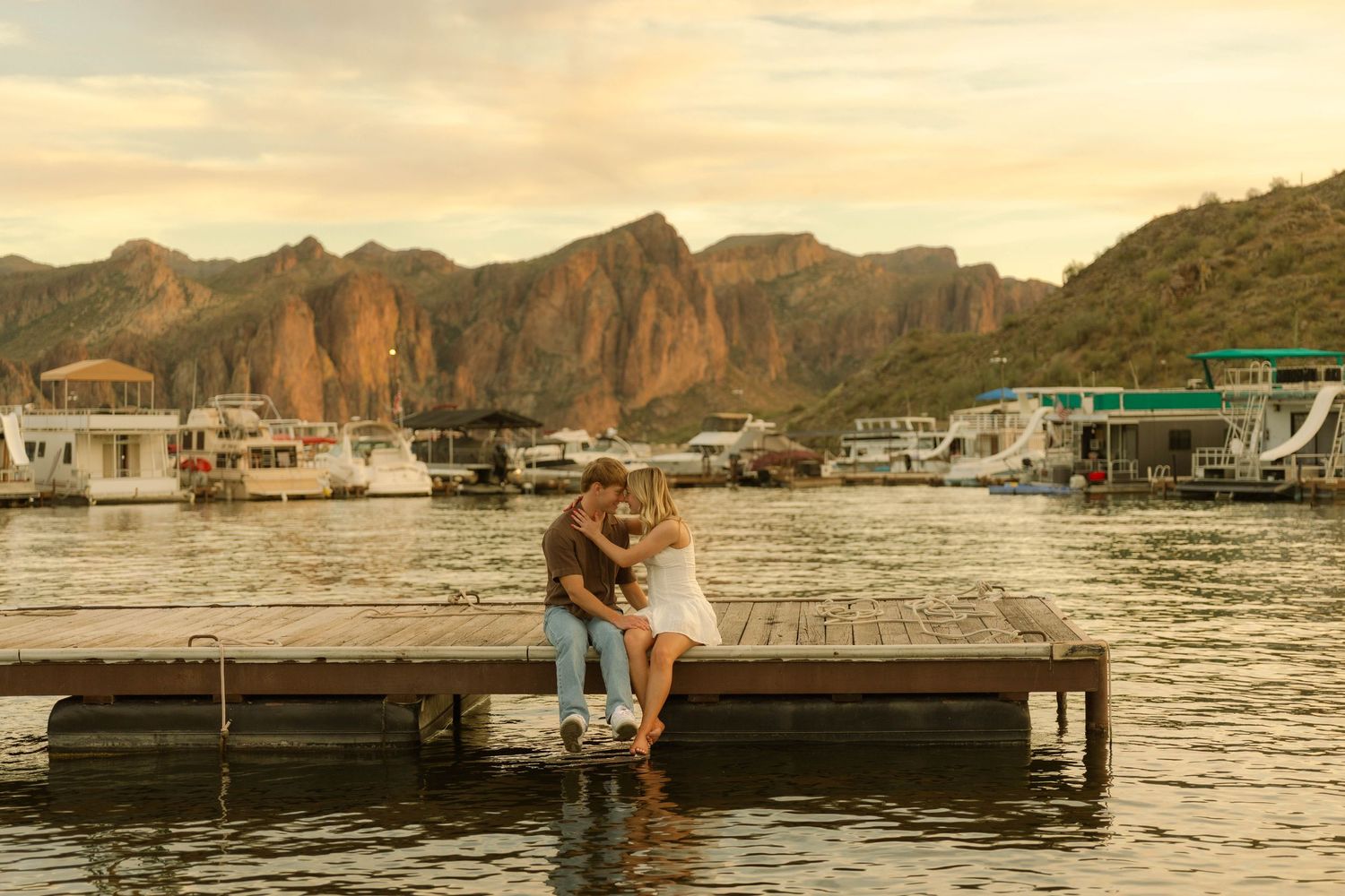 A couple embraces on a wooden dock at sunset with dramatic mountain peaks and houseboats in the background.
