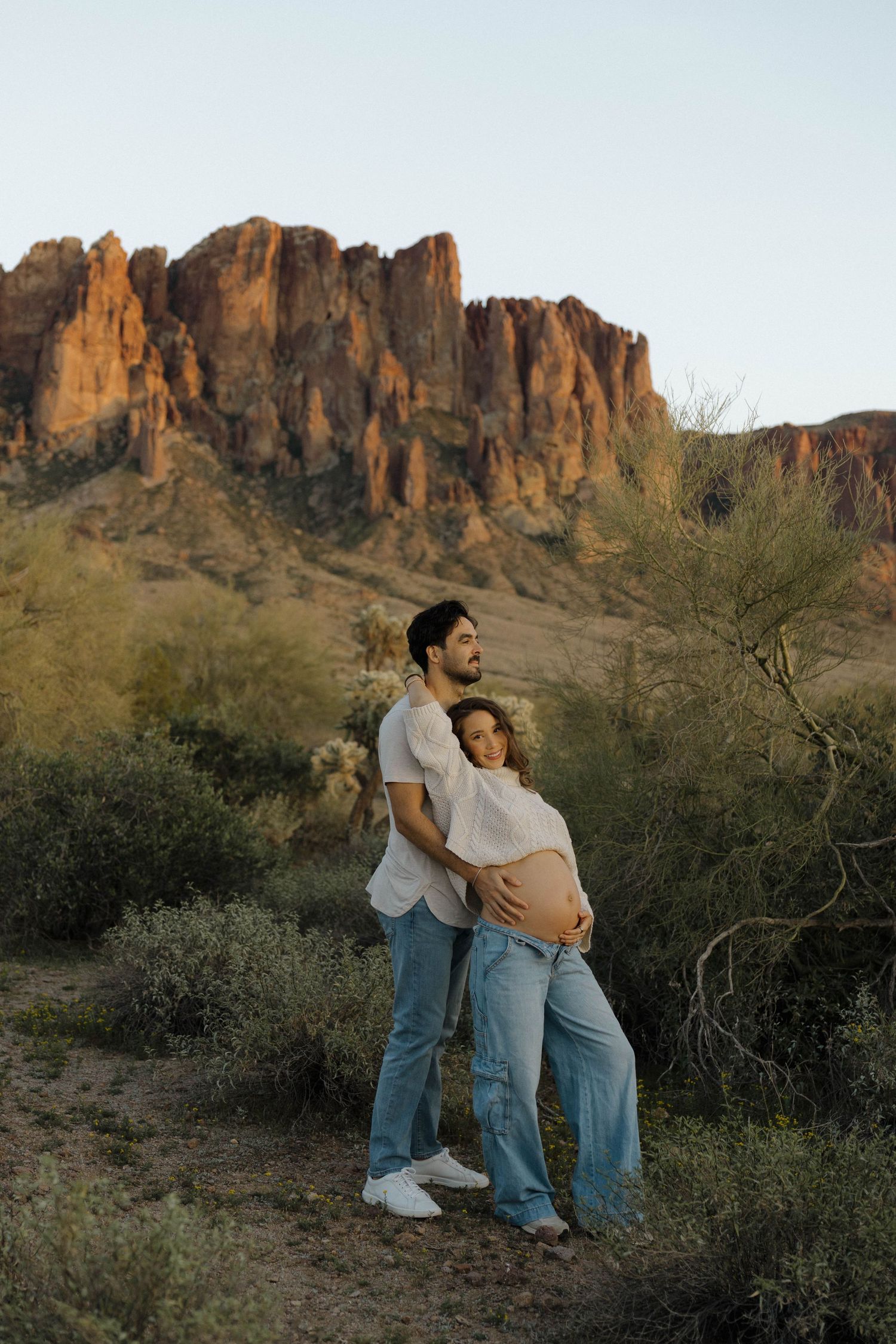 A couple embraces while standing among desert brush with rocky mountains in the background.