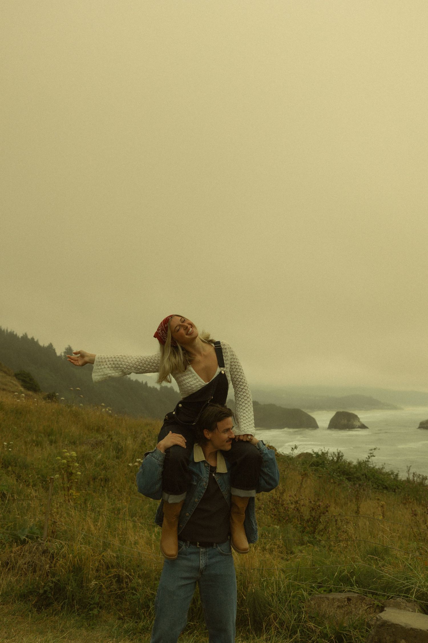 A woman sits on a man's shoulders on a misty coastal hillside, arms outstretched, with rocky ocean views behind them.