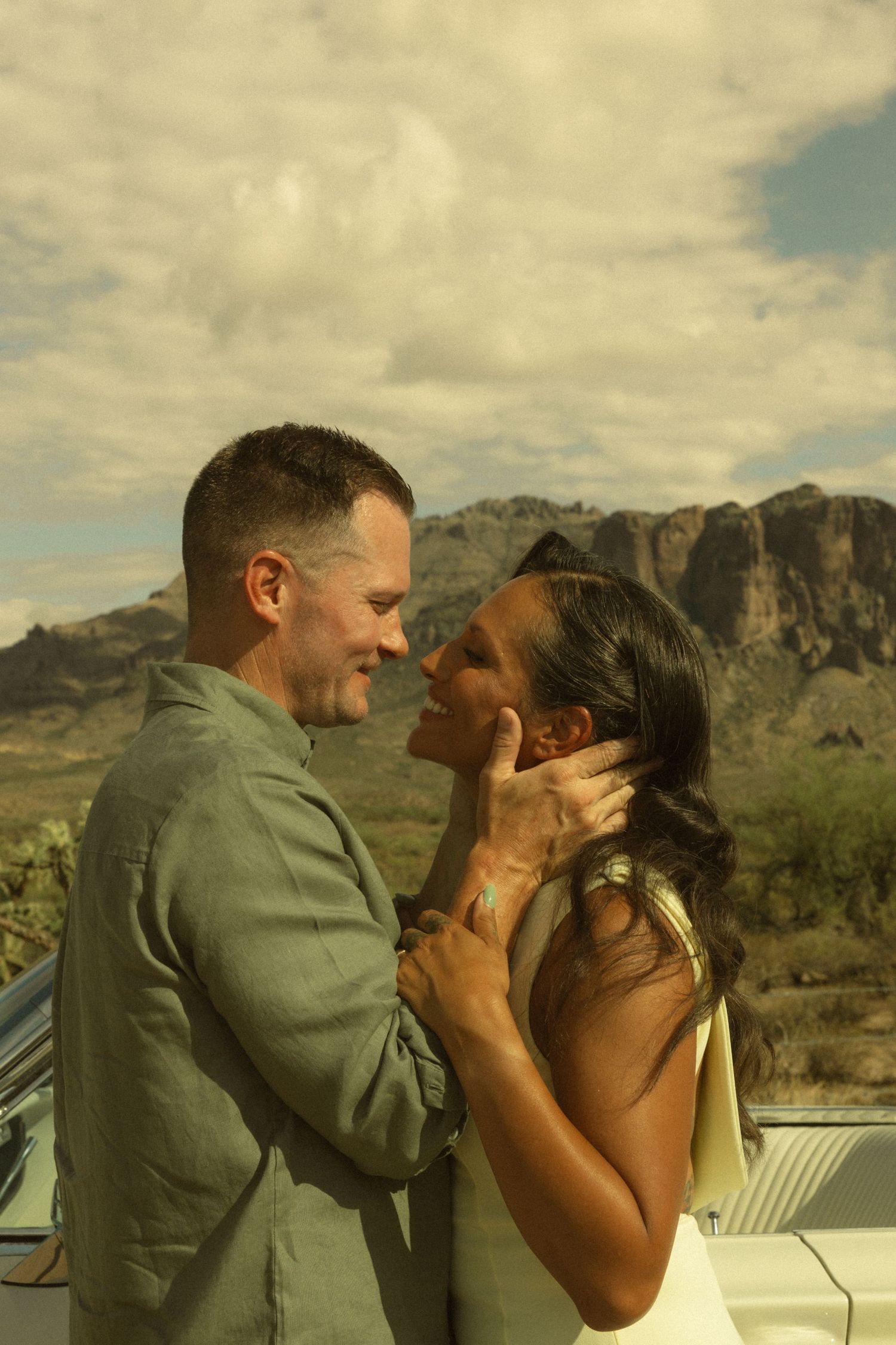 Couple sharing a romantic moment in the desert with mountains and cloudy sky backdrop near a vintage car.