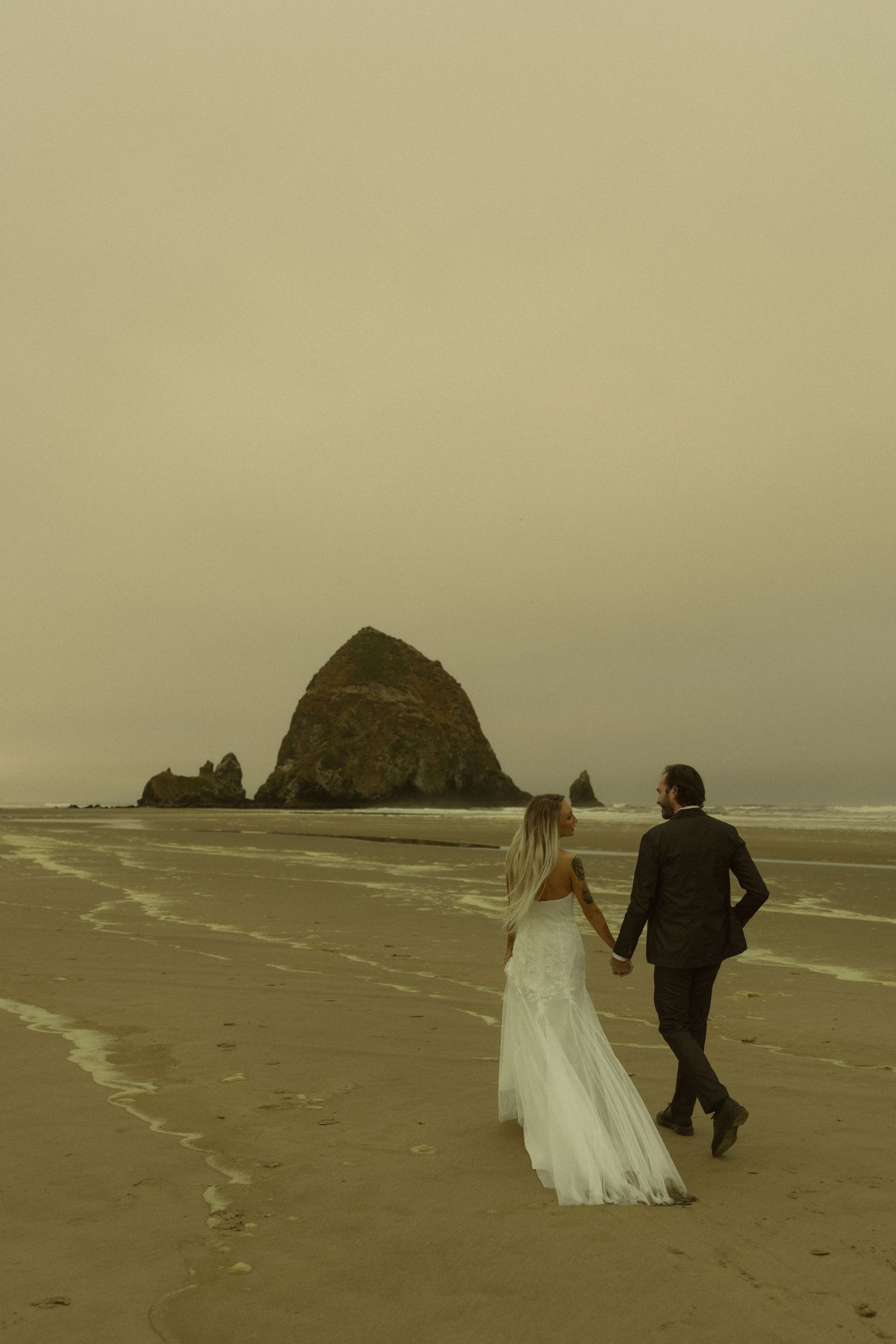 Bride and groom walk hand in hand on a misty beach with Haystack Rock at Cannon Beach, Oregon in the background.