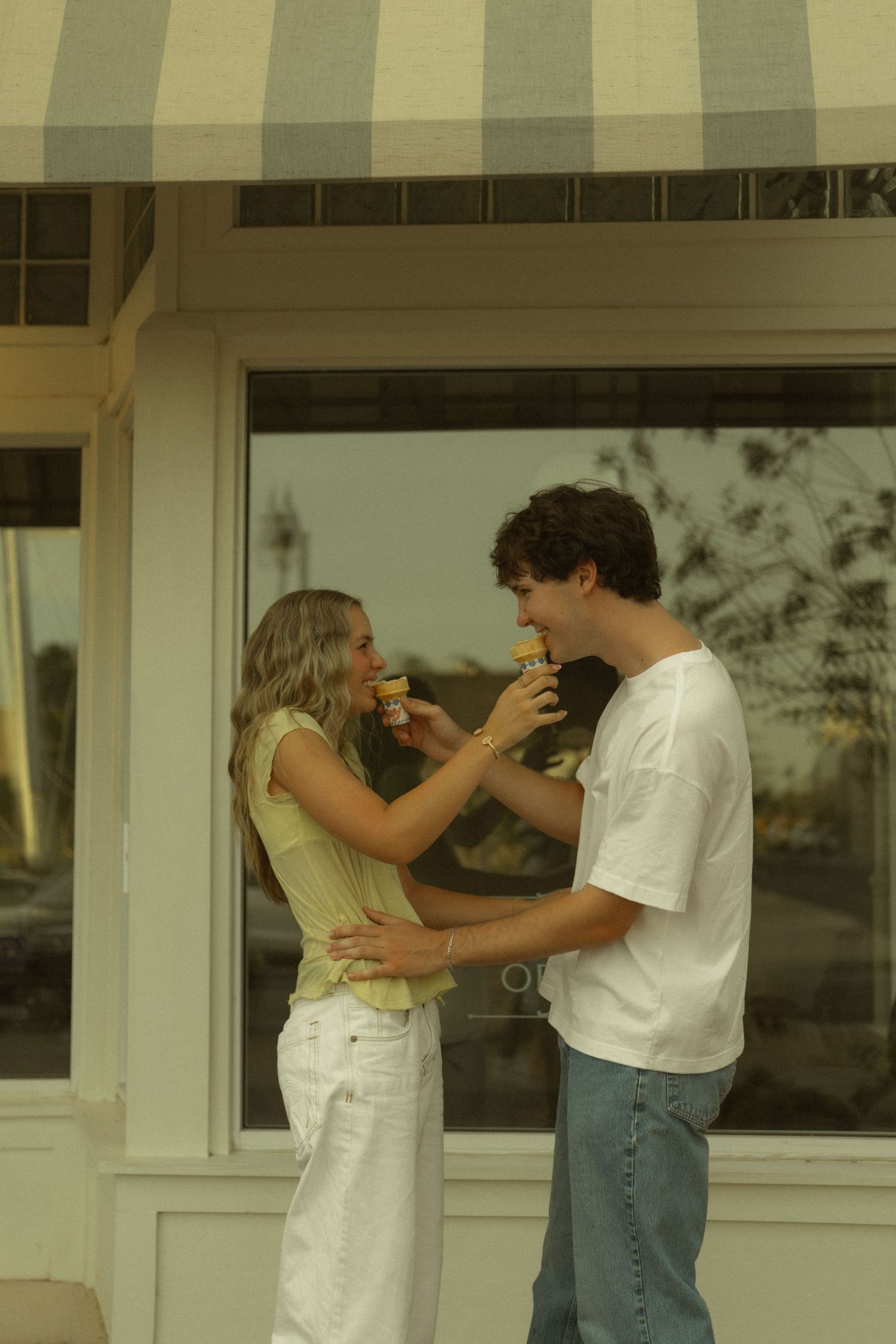 Young couple sharing ice cream cones outside a shop with striped awning on a warm summer day.