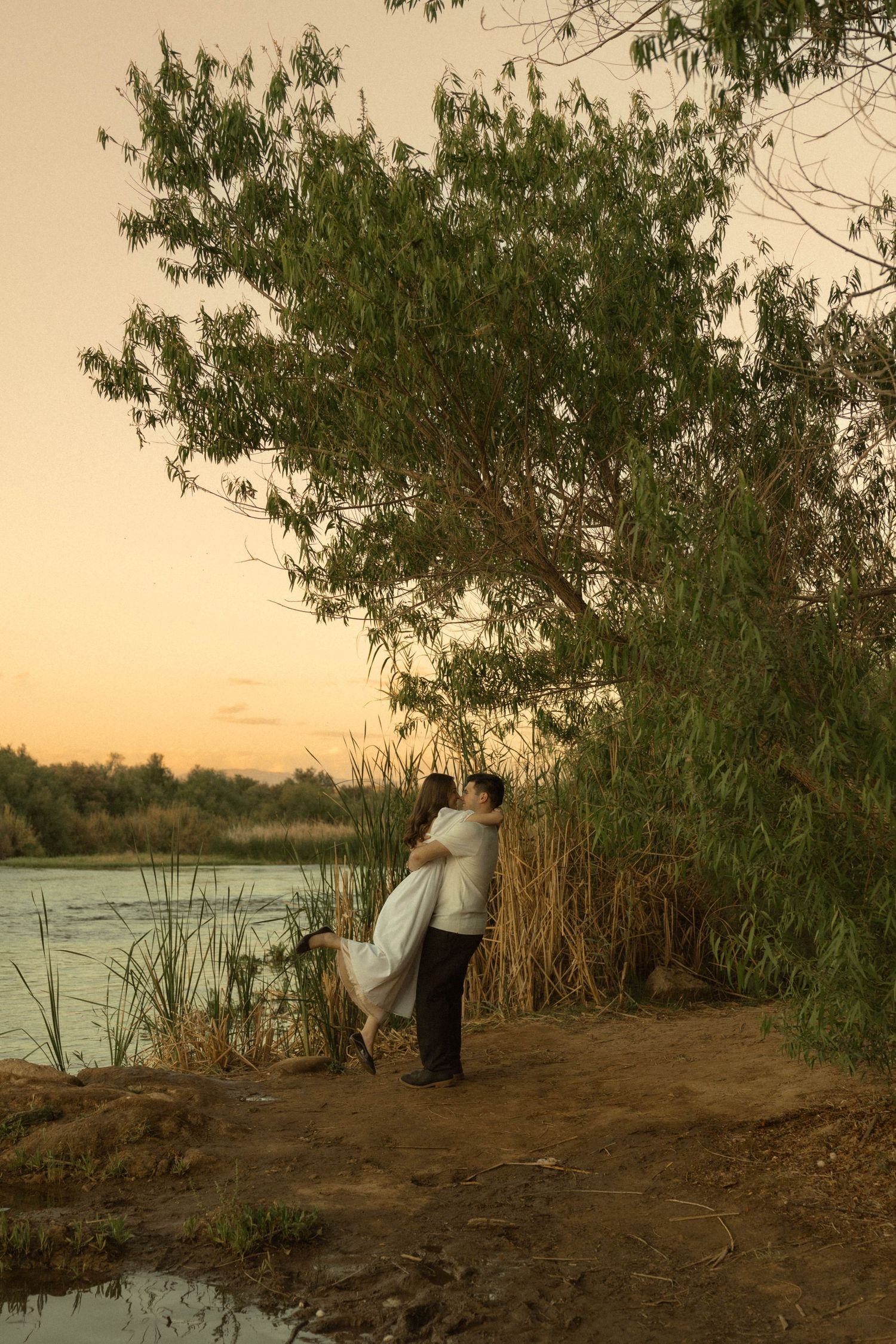 Couple sharing a romantic embrace near a large tree by a river at dusk, woman in flowing white dress.