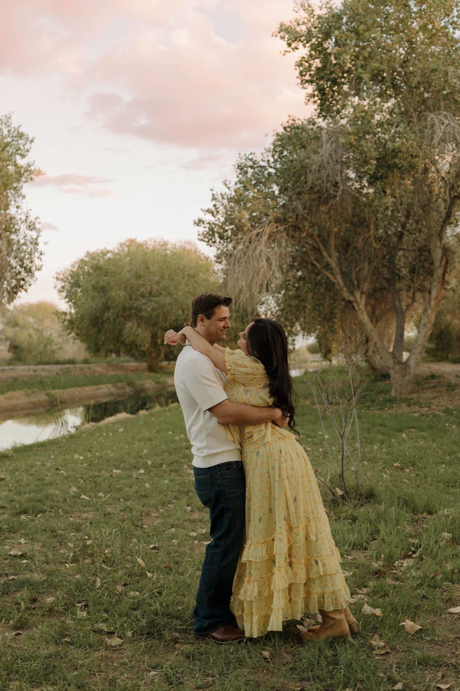 Couple dancing together on grassy hillside with trees and pink sky.