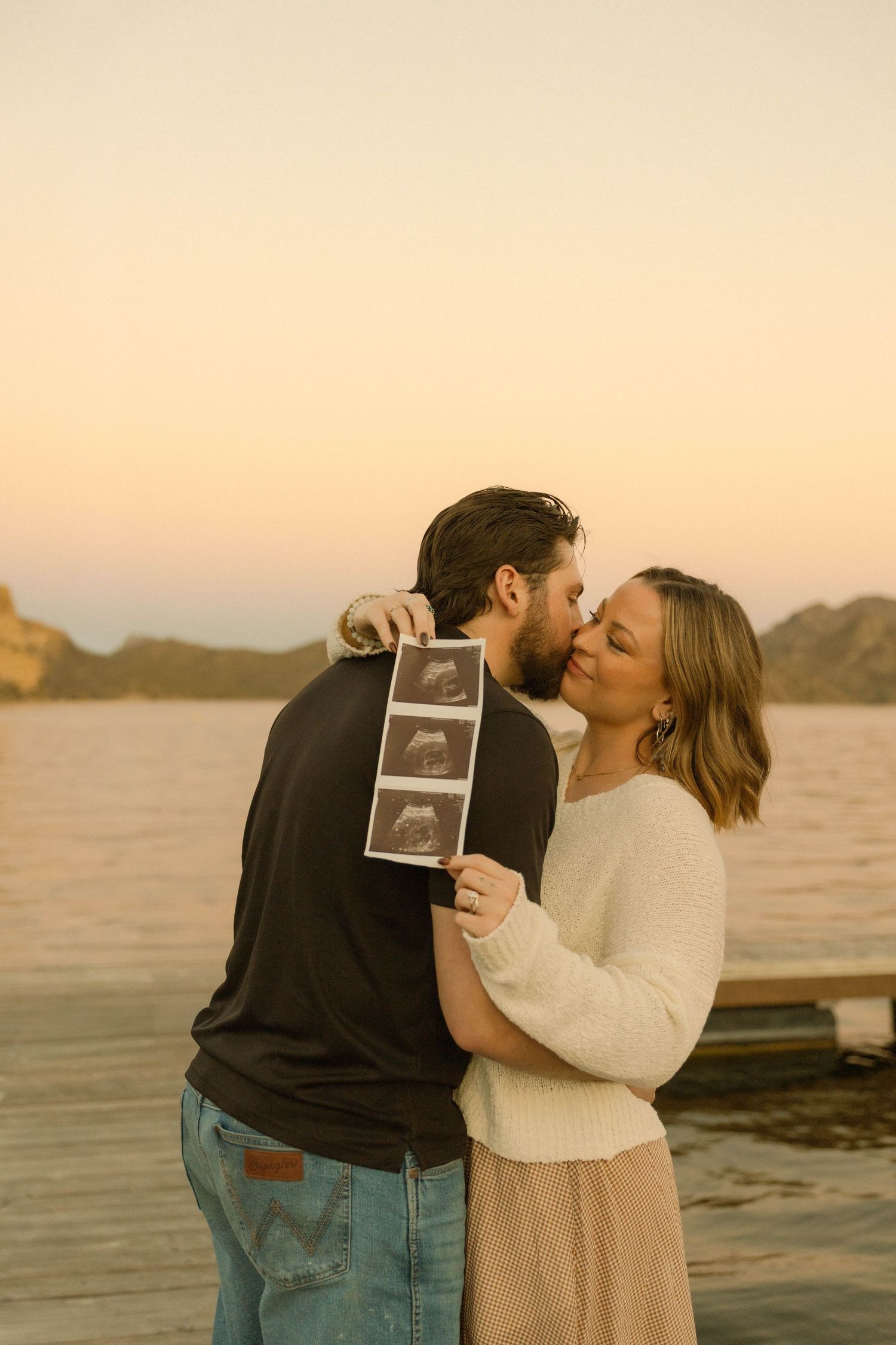 A couple shares a kiss while holding an ultrasound photo during sunset at a waterfront location.