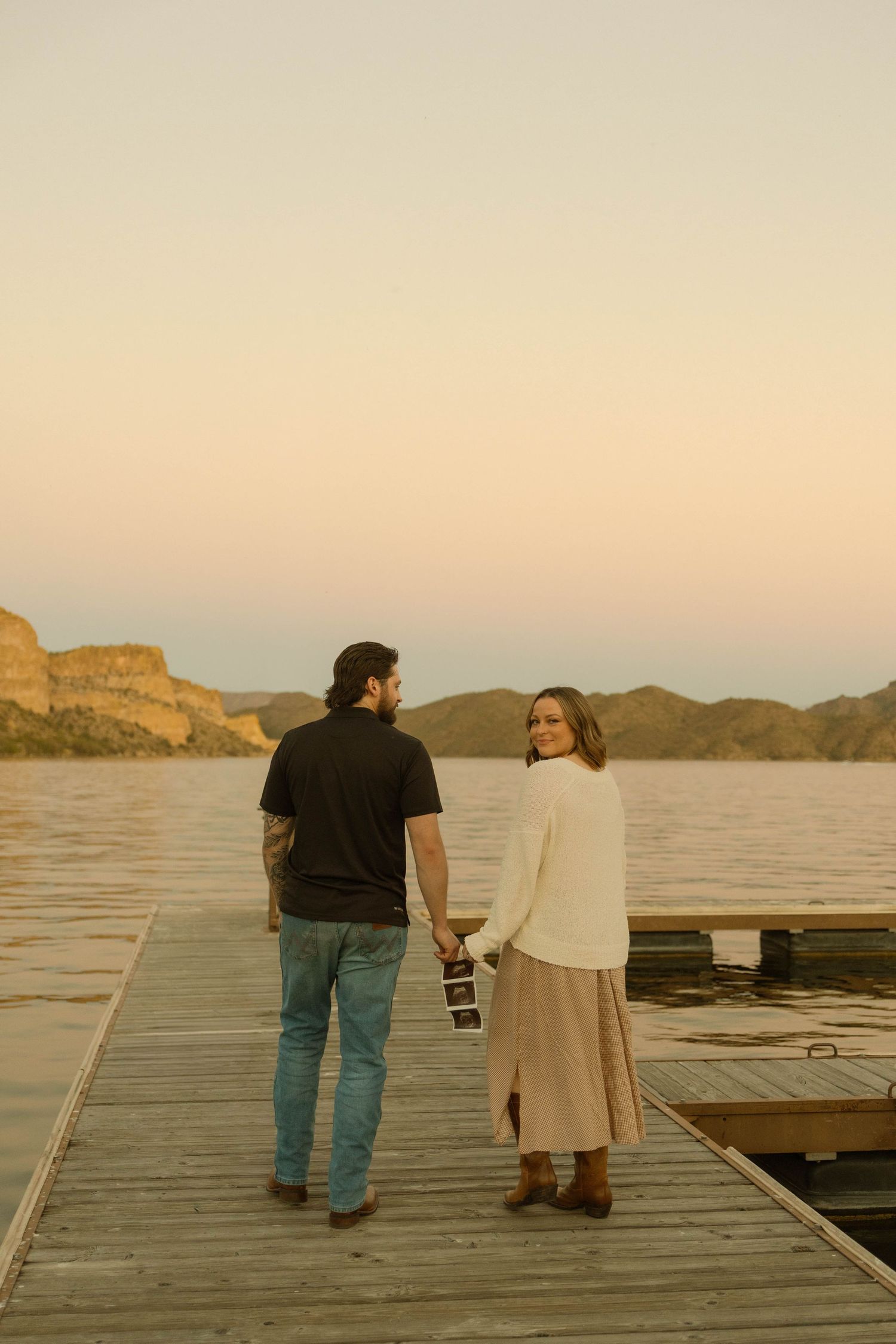 Two people stand together on a wooden dock overlooking a serene lake at sunset with mountains in the distance.