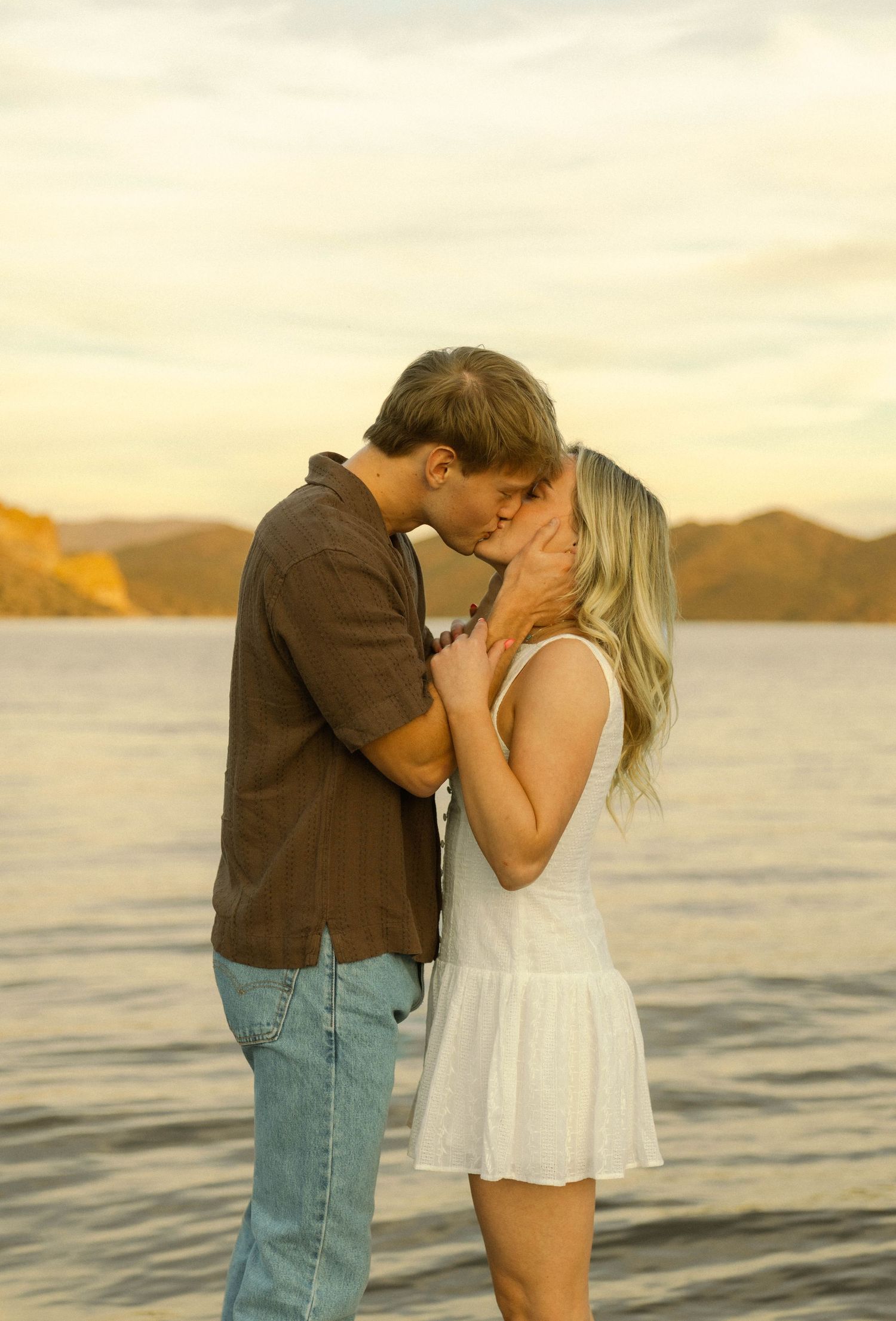 A romantic couple shares a tender moment at sunset by the water with mountains in the background.