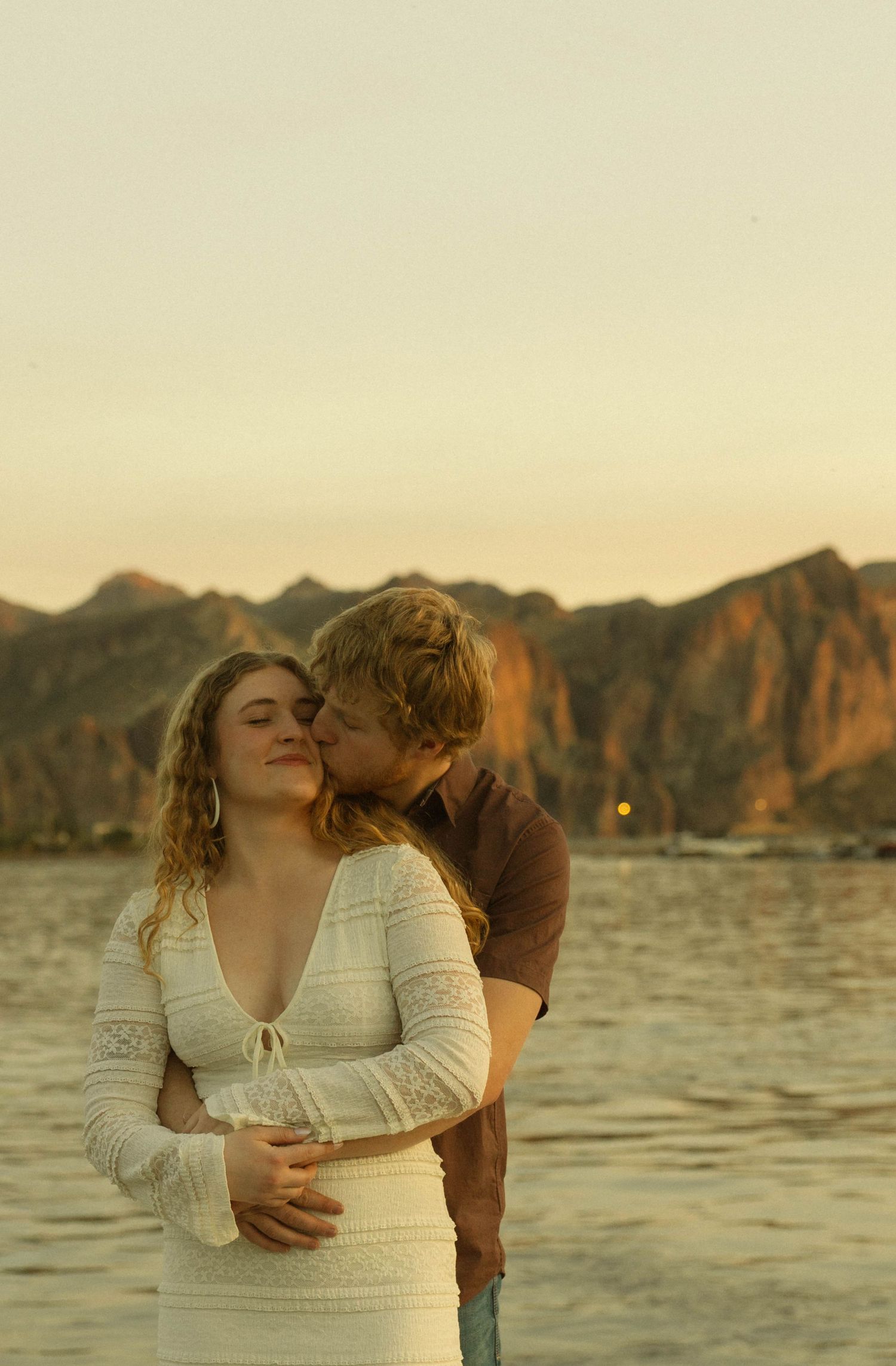 A romantic sunset moment by a mountainous lake with a couple in white dress and brown shirt embracing.