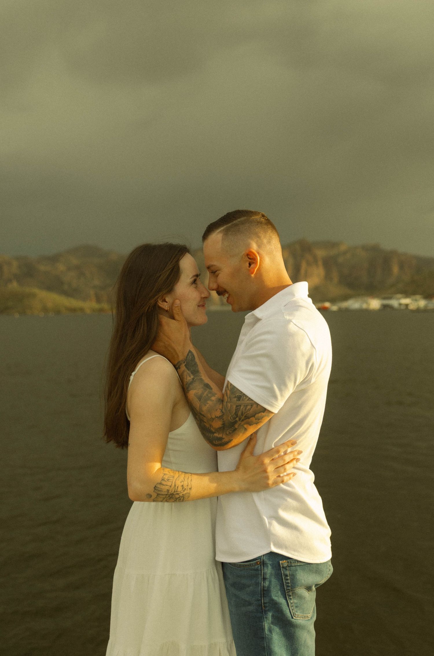 Romantic couple embraces against a scenic mountain backdrop at sunset near the ocean.