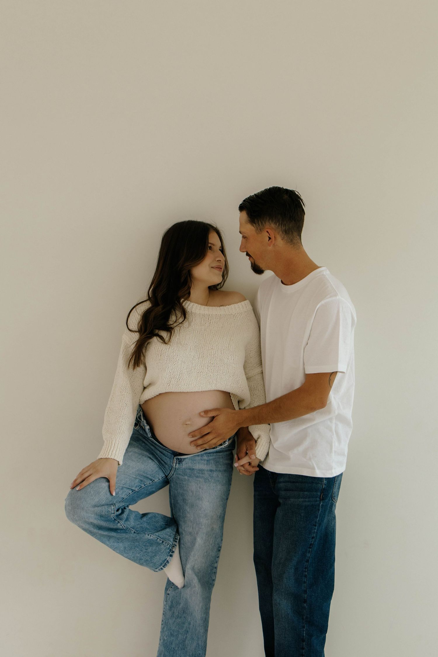 A couple in casual denim and white tops share intimate poses against a neutral wall in a minimalist photo series.
