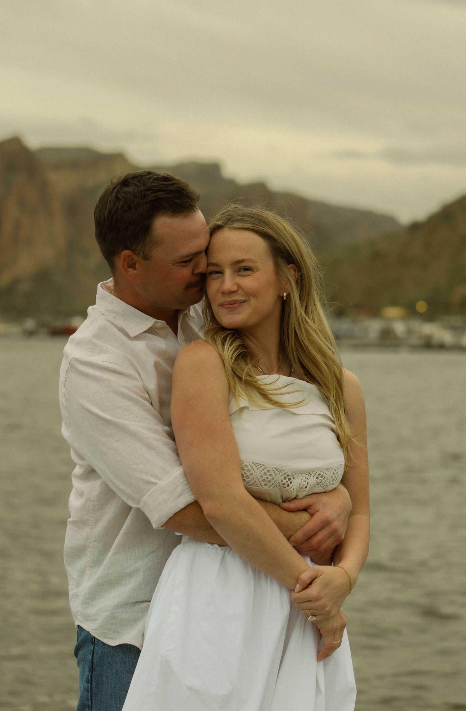 A tender embrace between a couple in white clothing against a mountainous waterfront landscape.