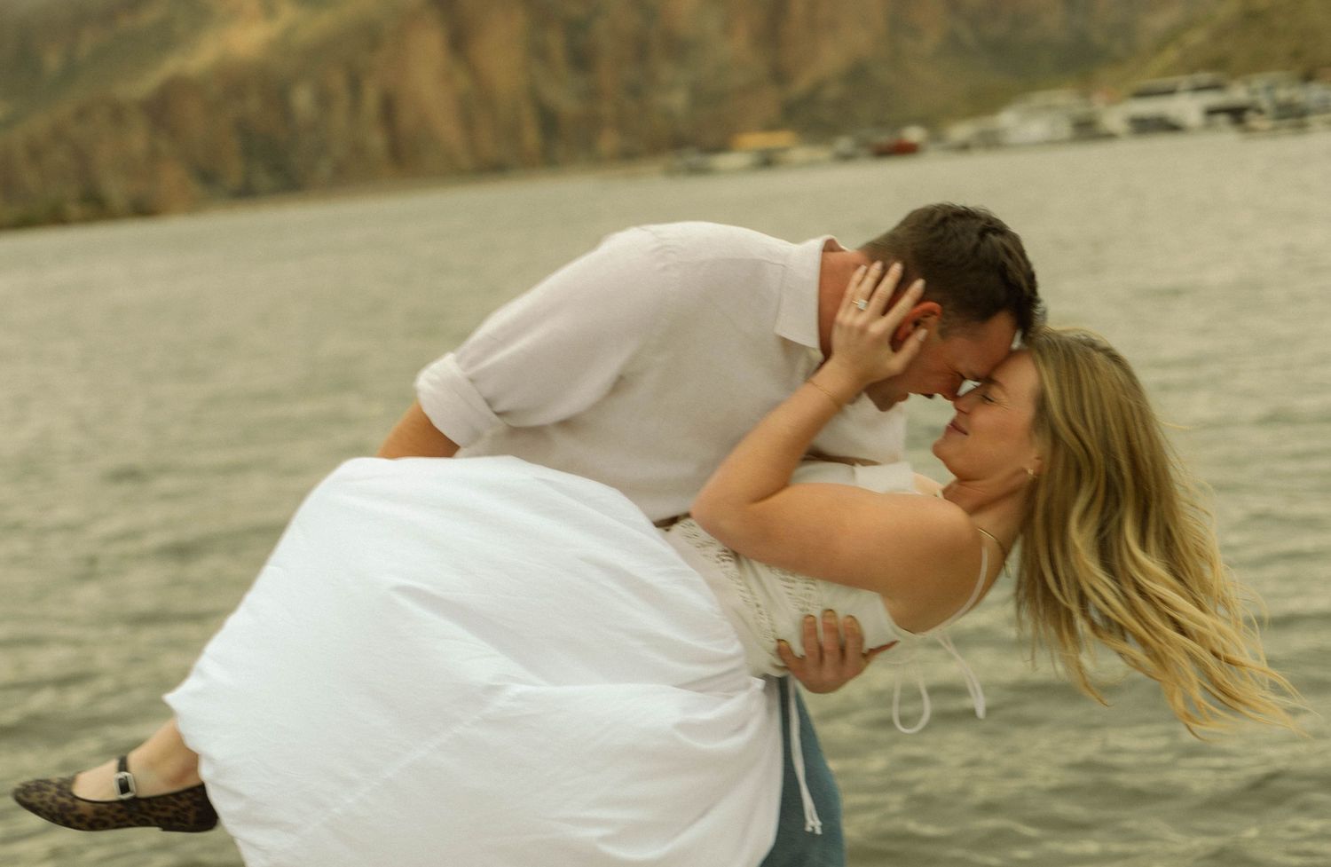 A couple embracing and kissing on a beach with mountains and water in the background during golden hour.