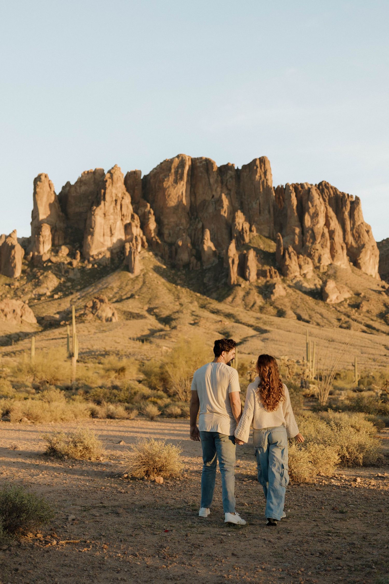 A couple walks hand in hand against the backdrop of majestic desert rock formations during golden hour.