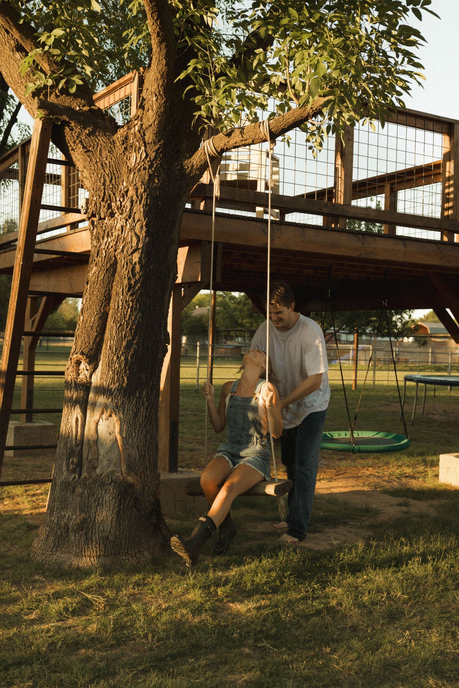 A man pushes a woman on a rope swing hanging from a large tree beside a wooden treehouse.