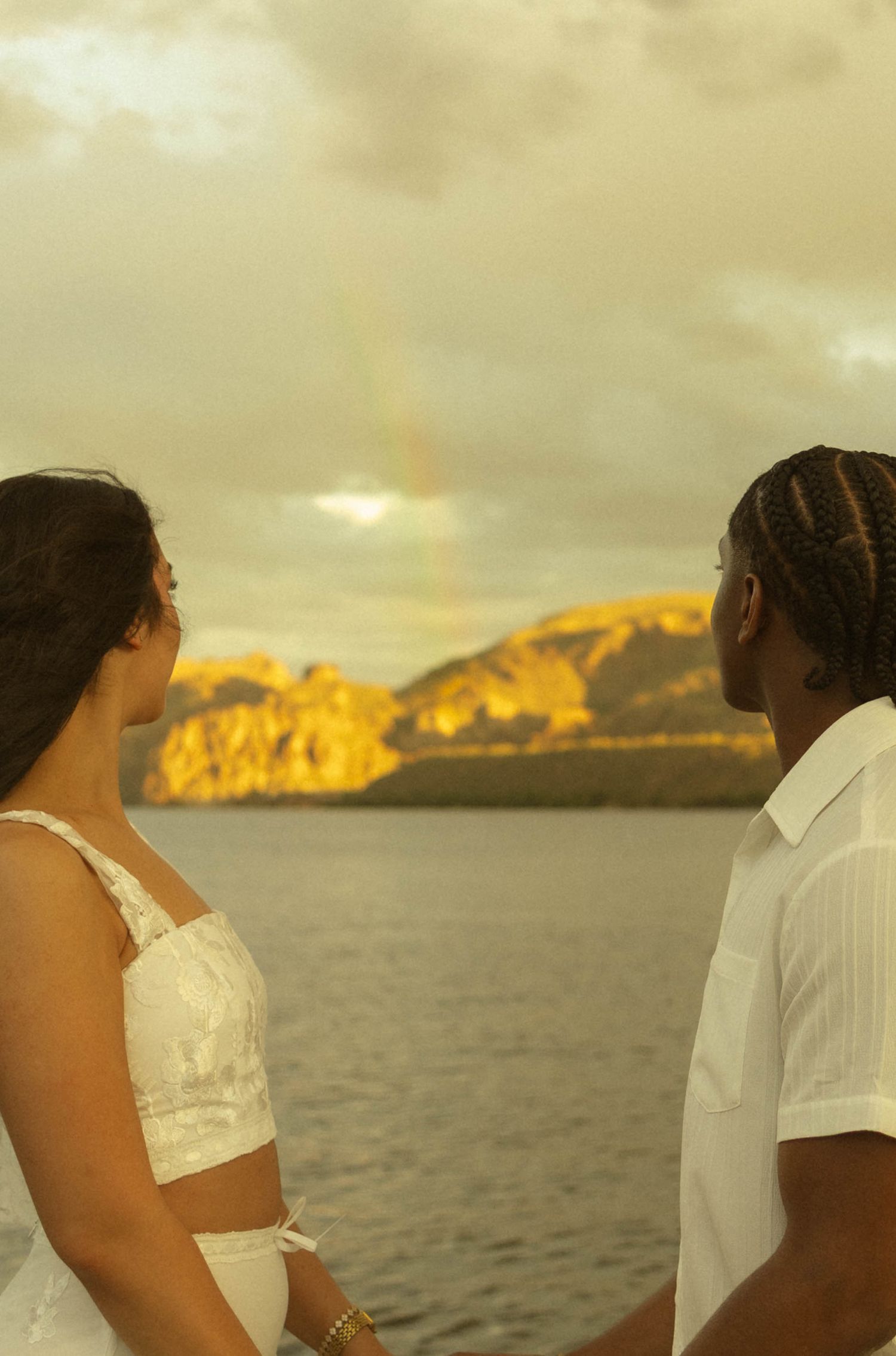 Couple in white outfits facing each other with a rainbow over golden-lit mountains and calm water at sunset.