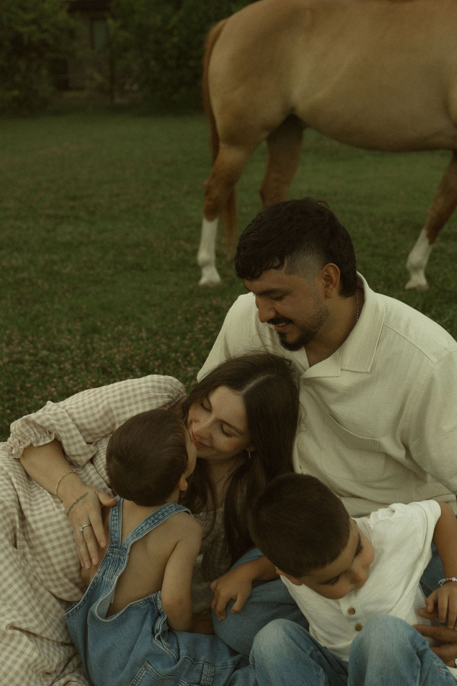 Family of four sitting together on grass laughing with a horse in the background on a sunny day.