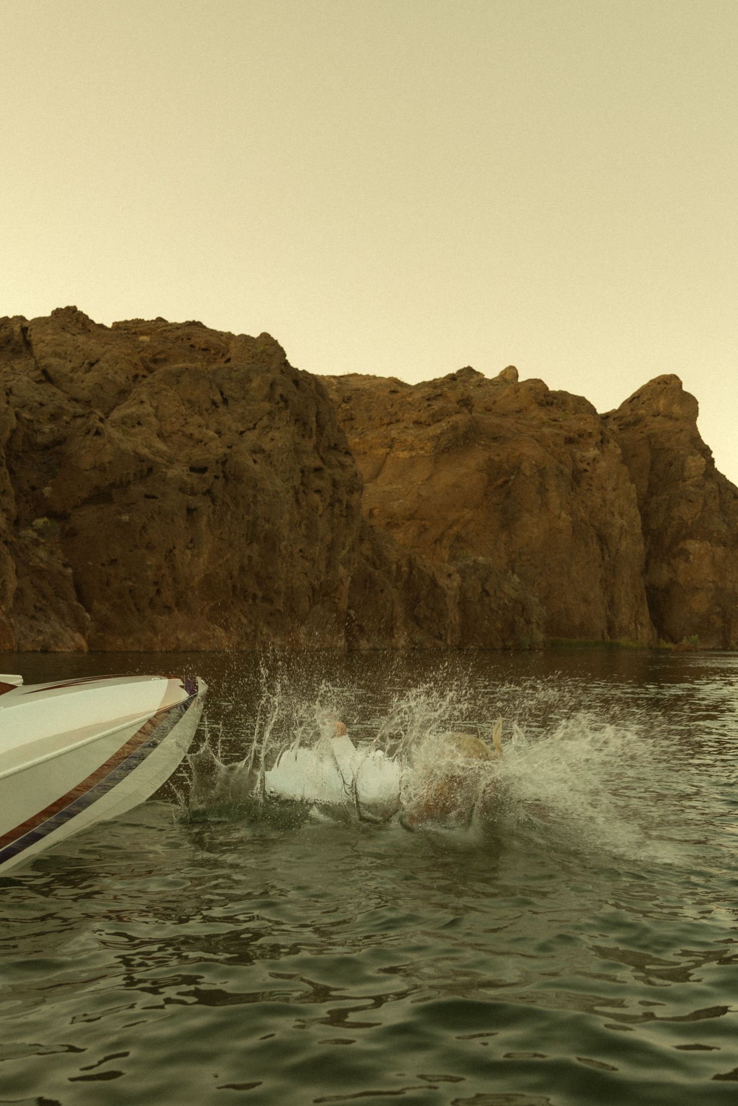A speedboat creates dramatic water splashes near rugged brown rocky cliffs on a calm lake at dusk.