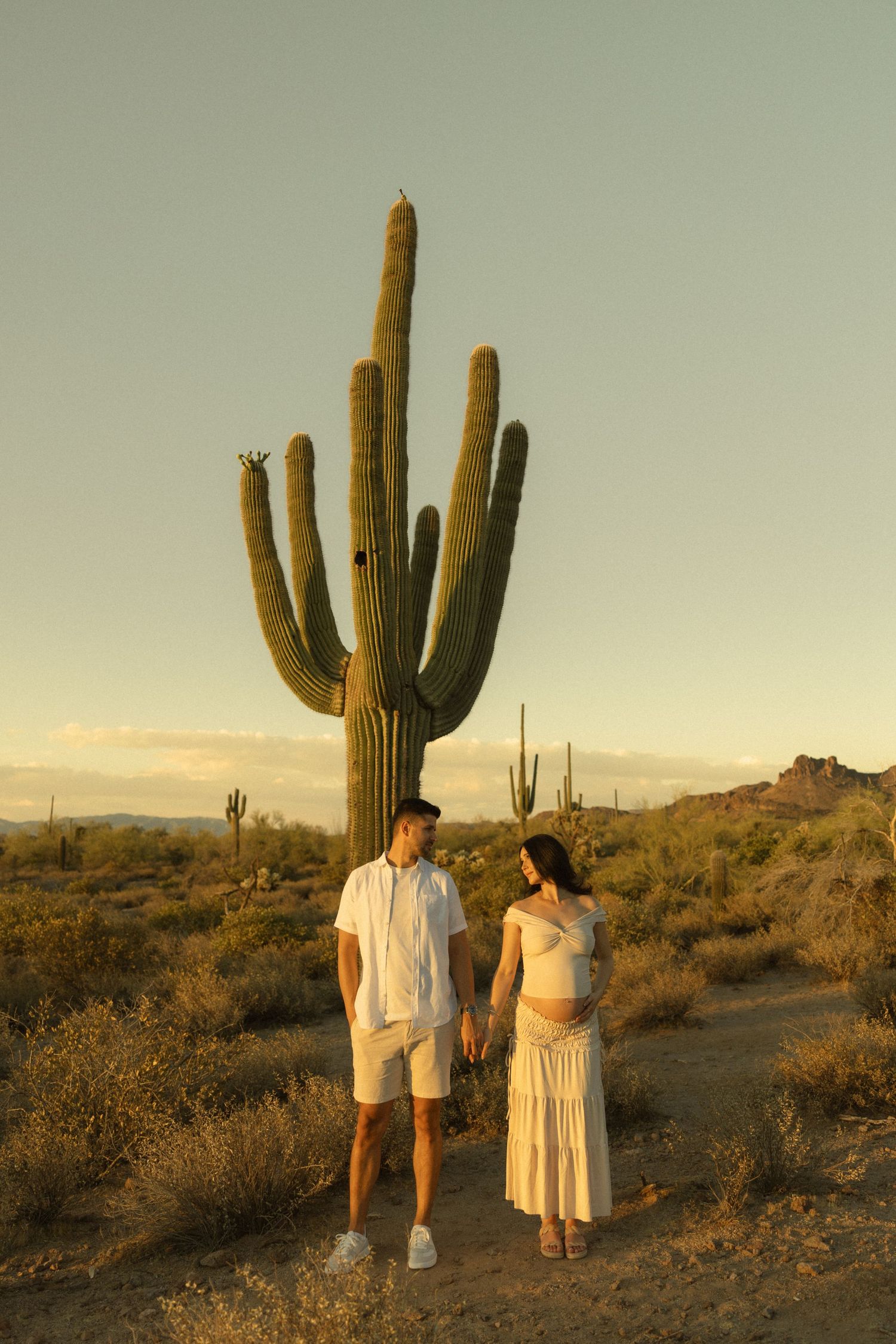 Pregnant couple holding hands in Arizona desert at golden hour with tall saguaro cactus backdrop.