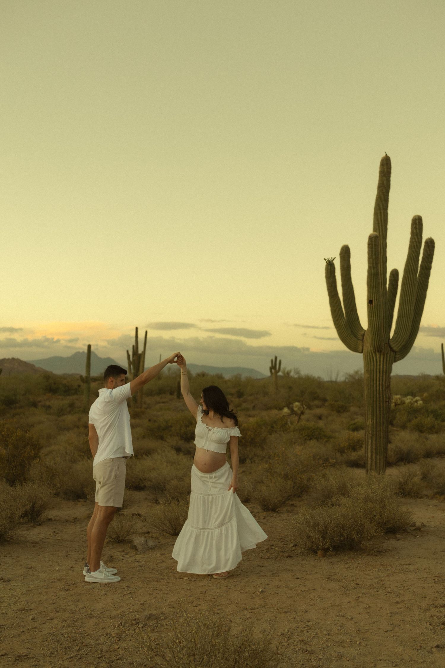 Pregnant woman in white dress being twirled by partner in Arizona desert at golden sunset hour.