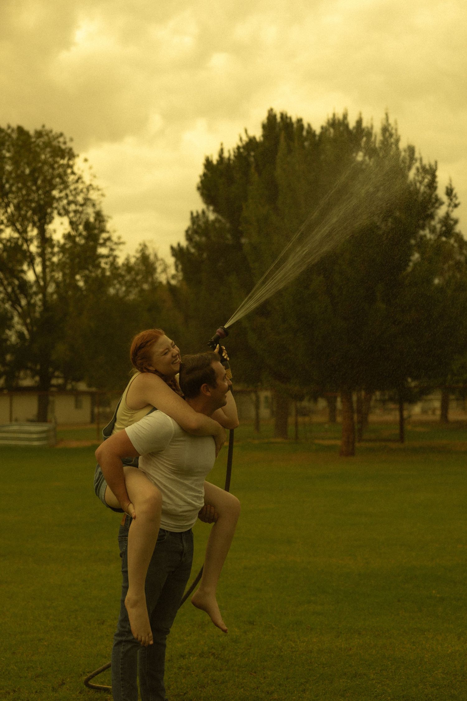 Man carrying woman on his back while she sprays a garden hose outdoors on a cloudy day.