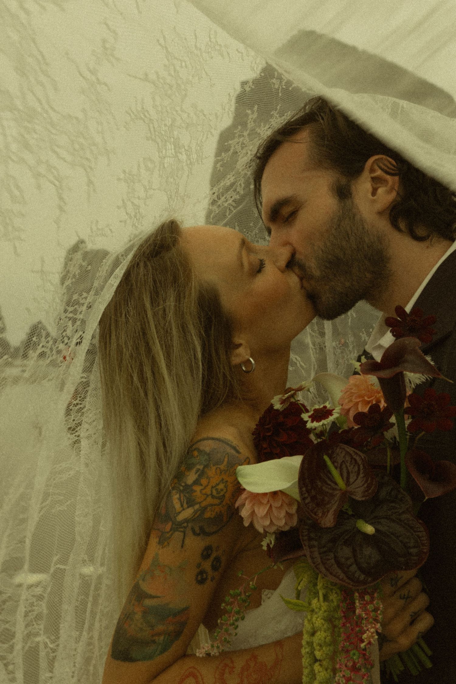 Tattooed bride and groom share a passionate kiss under a lace veil, bride holding a rich burgundy wedding bouquet.