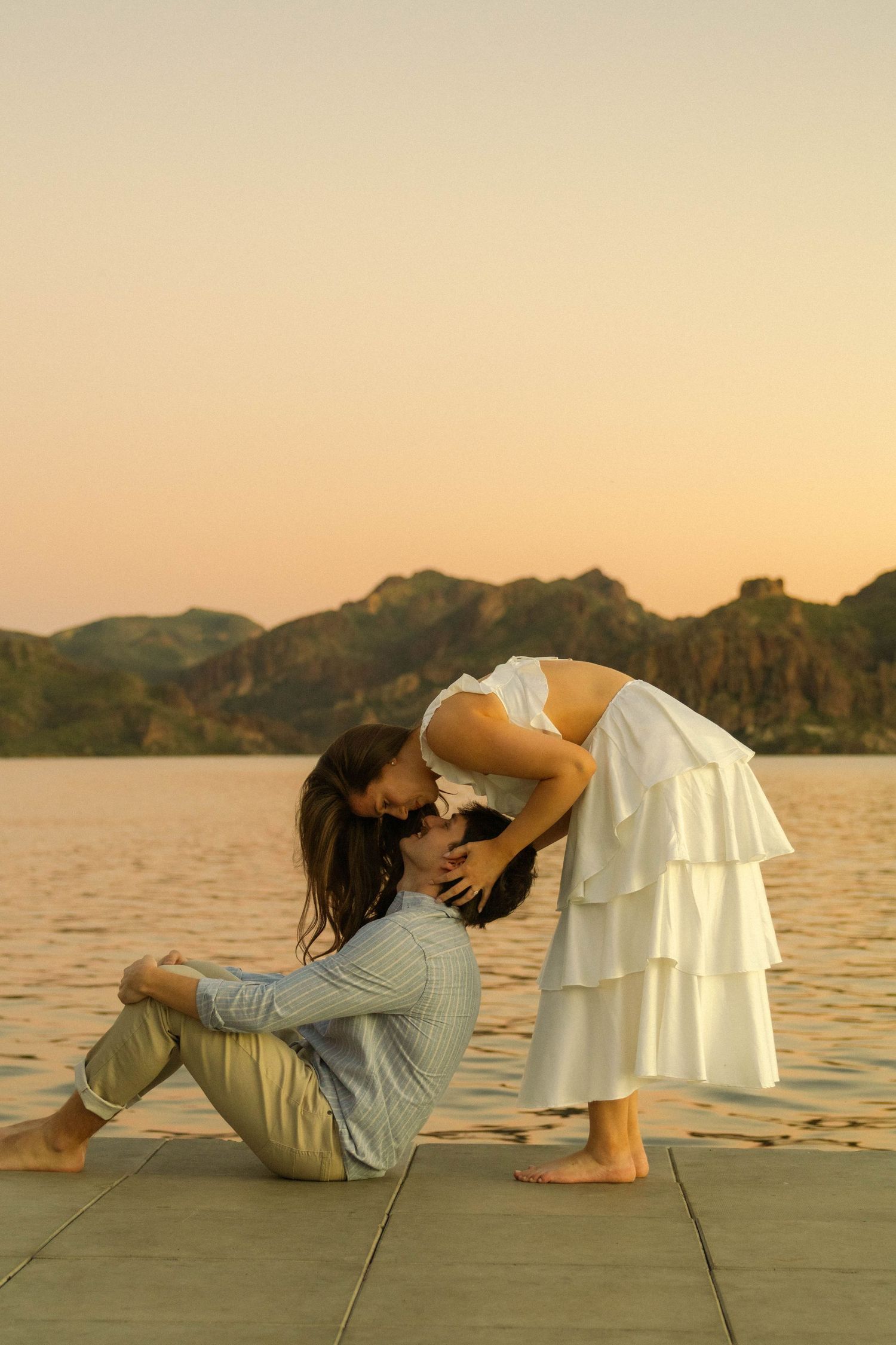 A couple reclines on a wooden dock during sunset with mountains and water in the background.
