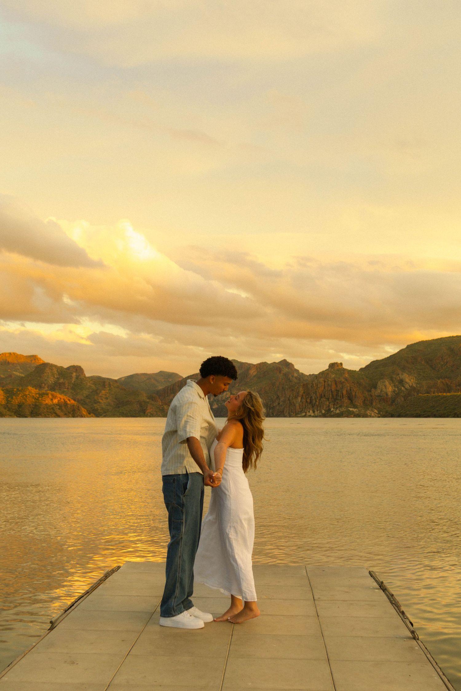 A couple shares an intimate moment on a wooden dock at sunset with mountains and water creating a romantic backdrop.