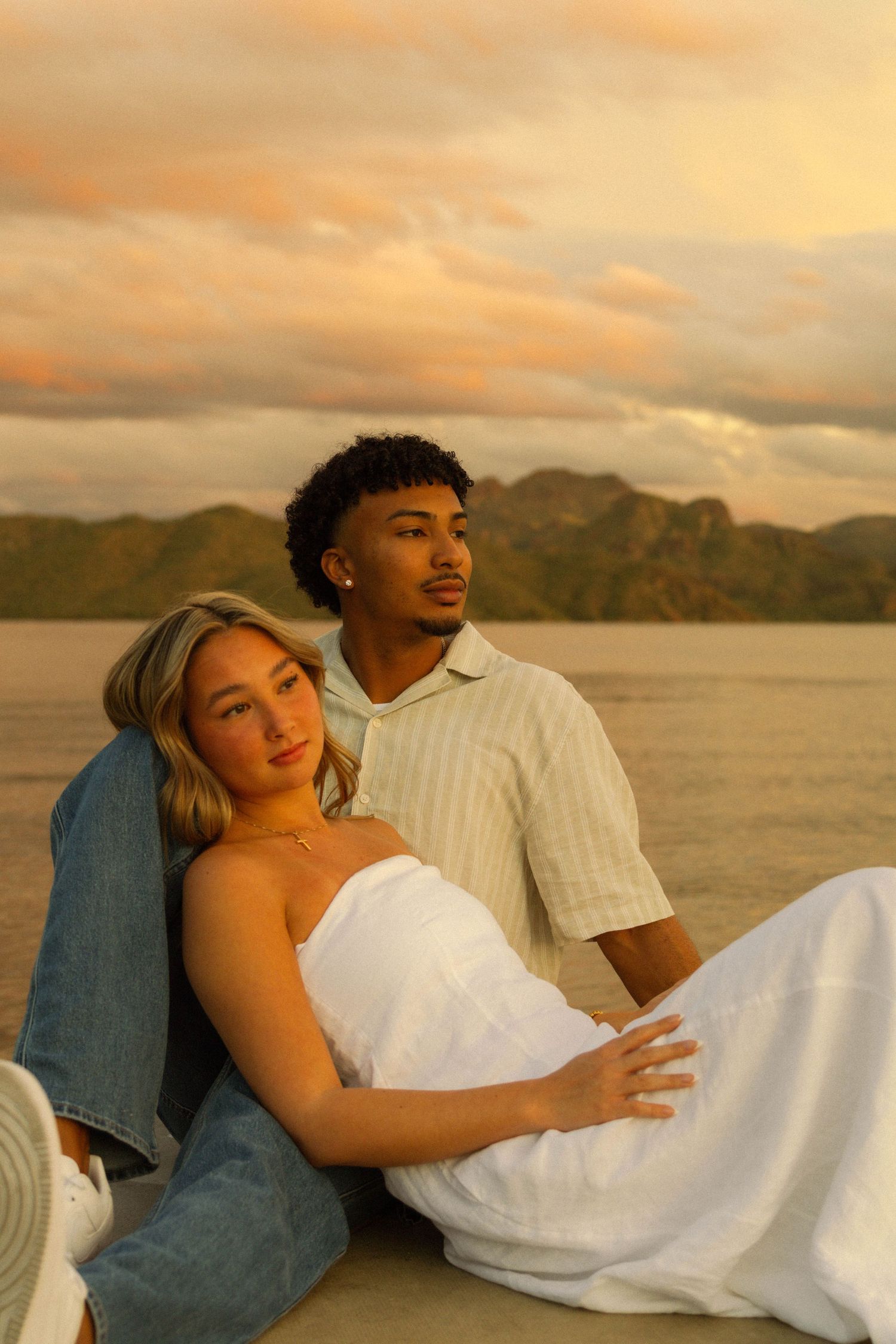 A couple sits together at sunset by a lake with mountains in the background, creating a romantic golden hour moment.