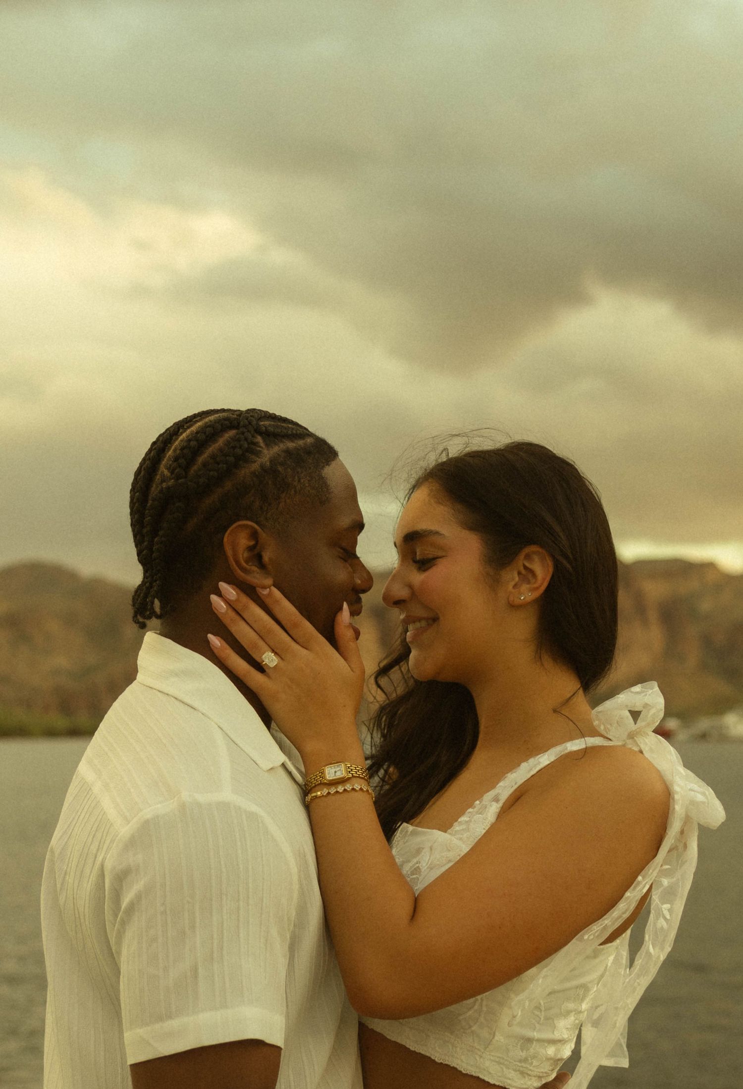 Romantic couple in white almost kissing by a lake with mountains and cloudy sky at sunset.