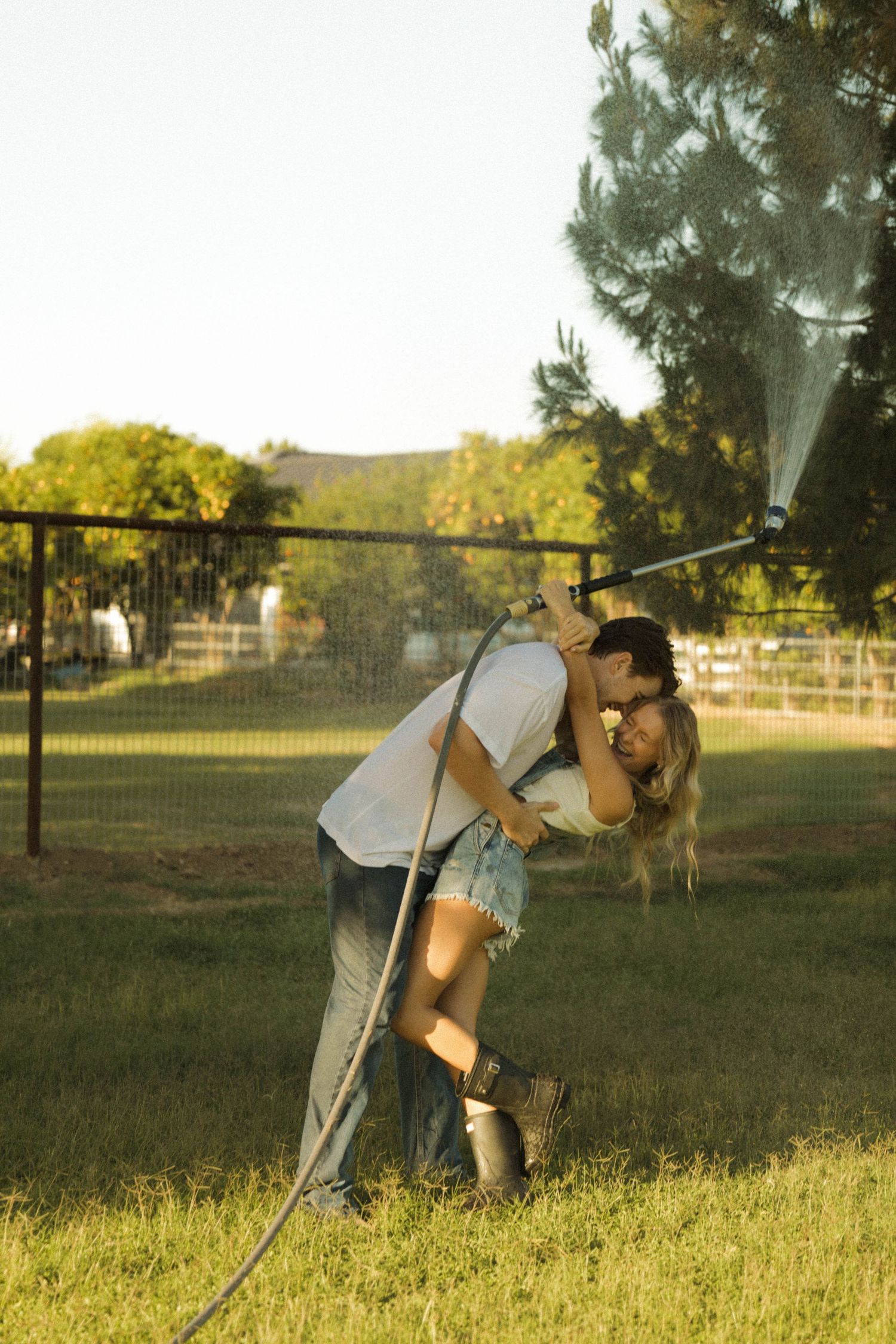 Romantic couple kissing while holding a garden hose spraying water in a sunny backyard.