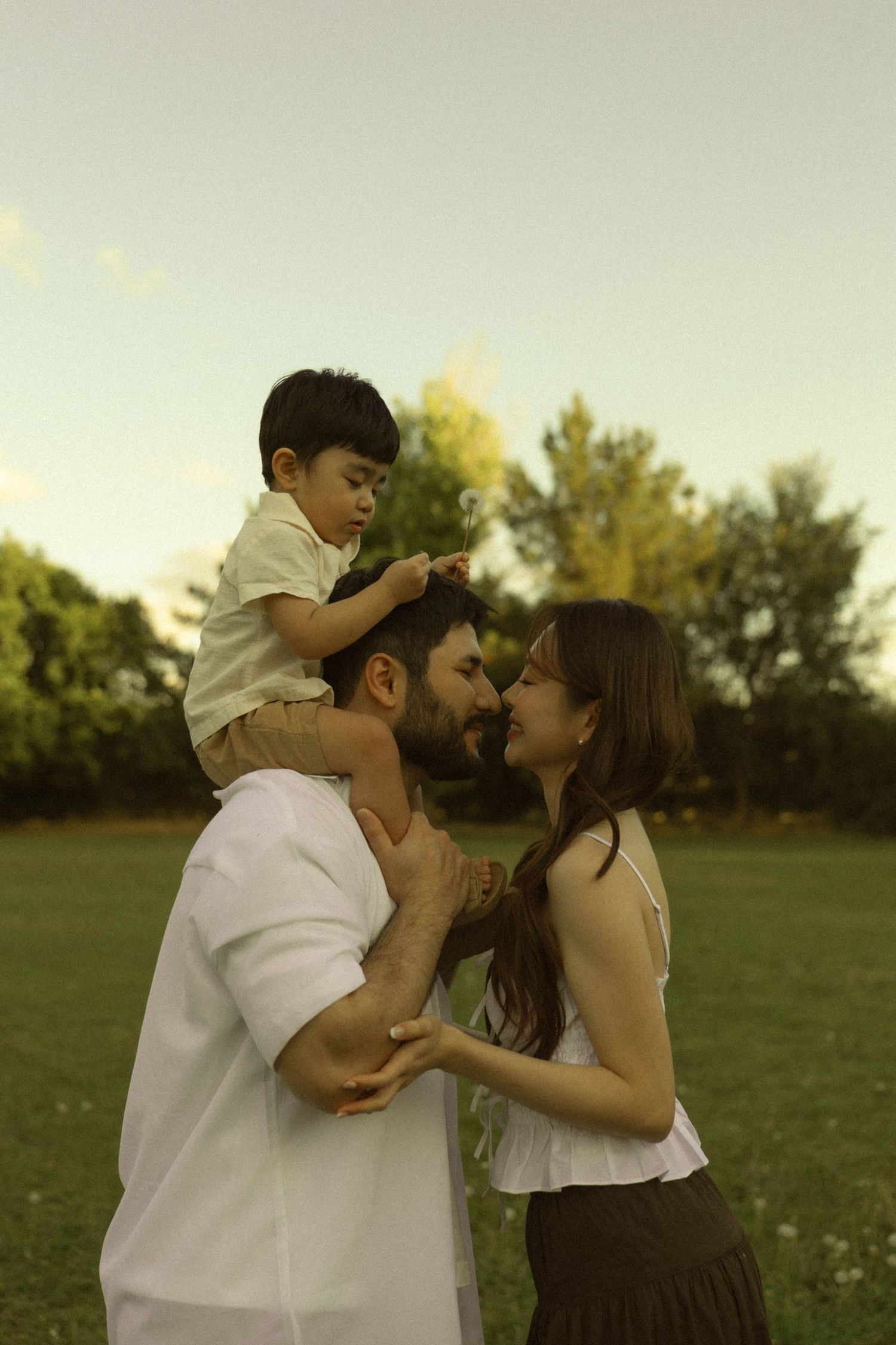 A bearded father carries his toddler son on his shoulders while his smiling wife holds his hands in a lush green park.