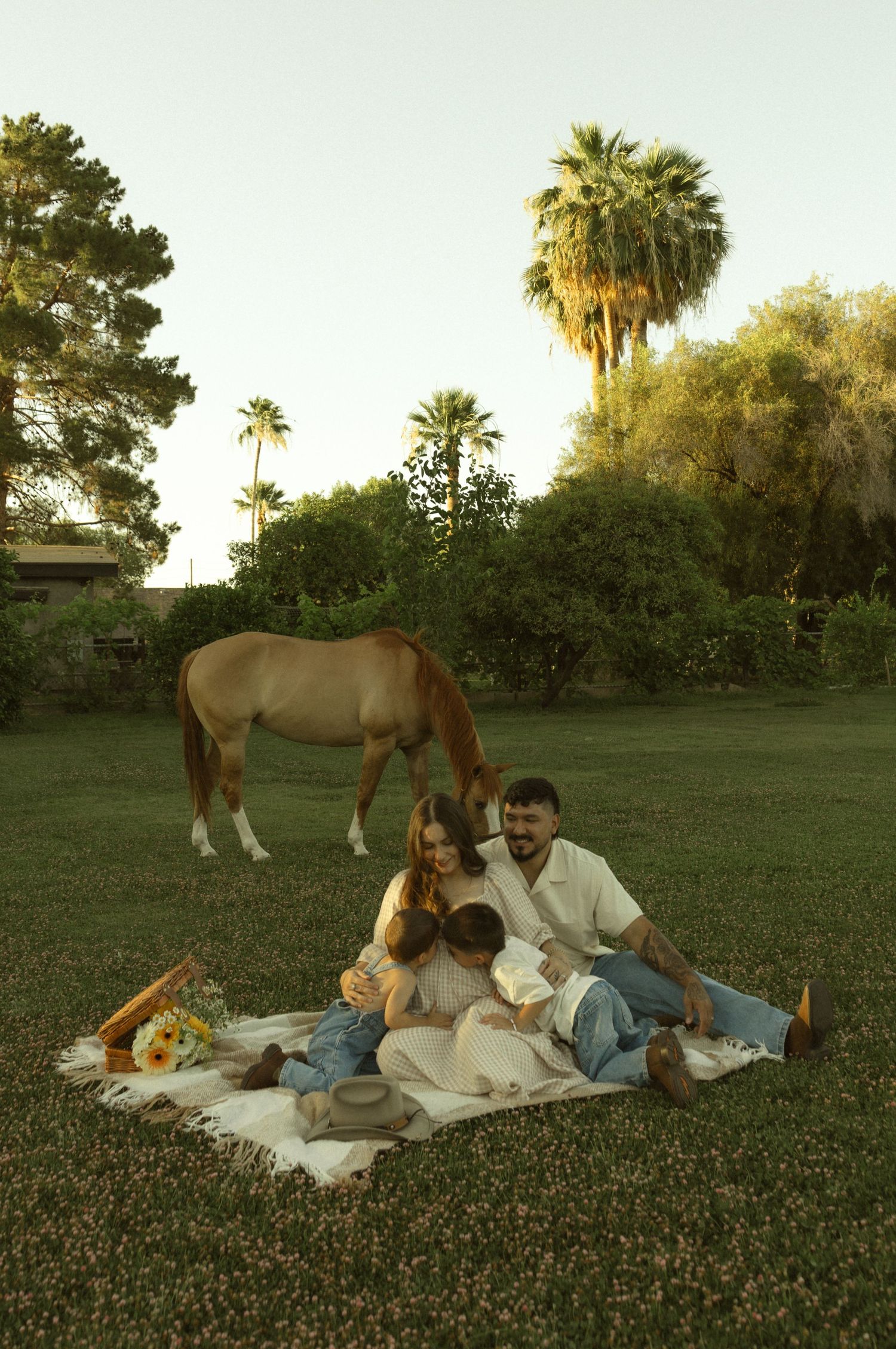 Family sitting on a blanket in a lush green yard with a grazing horse and palm trees in the background.