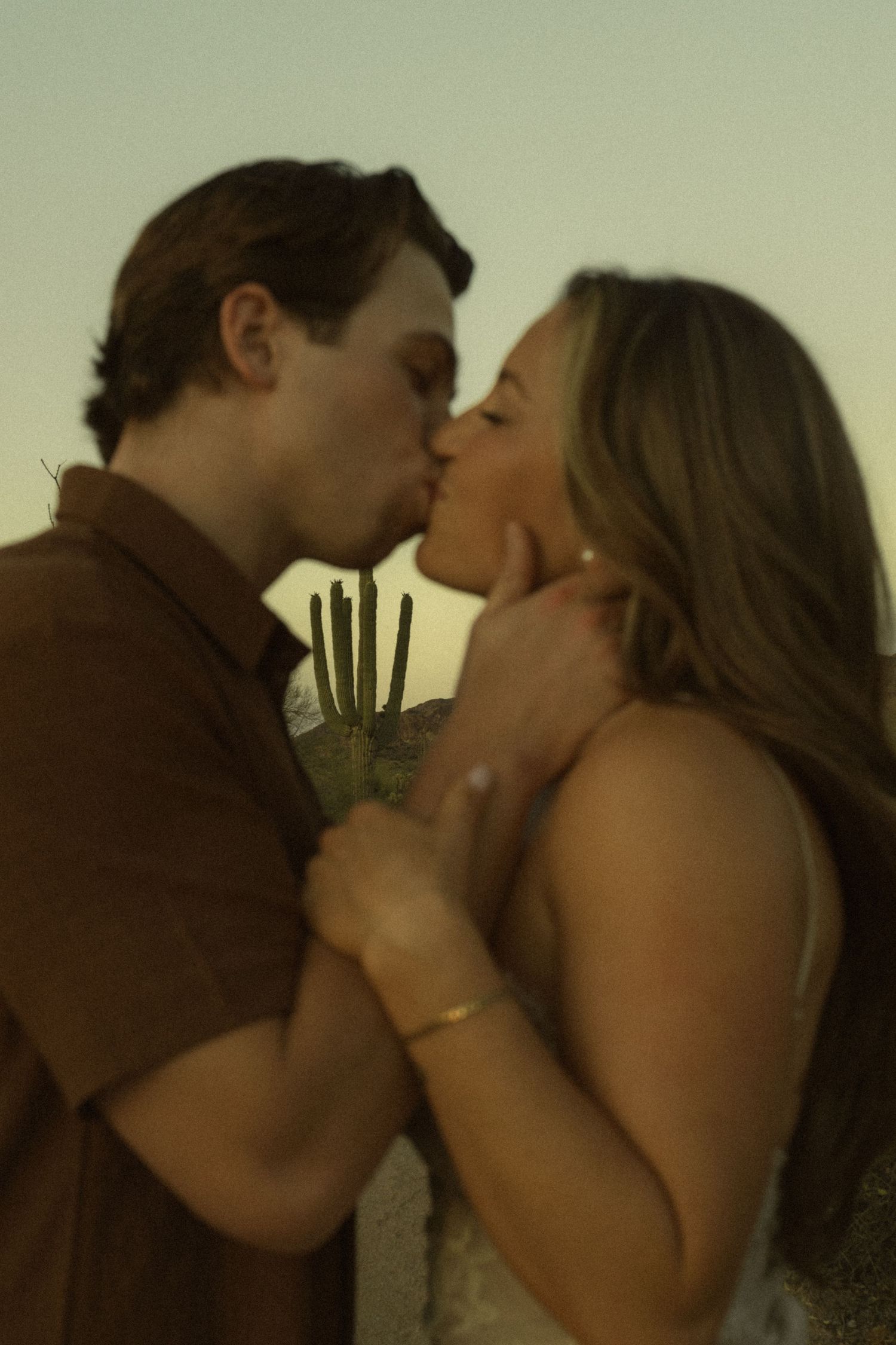 Couple sharing a romantic kiss at sunset with a saguaro cactus in the background.