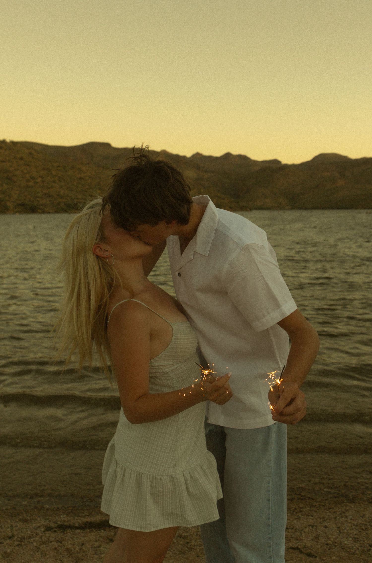 A couple shares a romantic kiss at sunset by a lake, holding sparklers, dressed in white summer outfits.