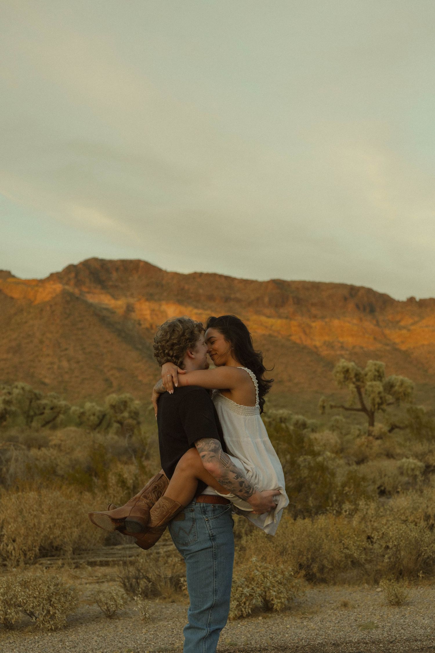 Couple sharing a romantic moment at golden hour with desert mountains and Joshua trees in the background.