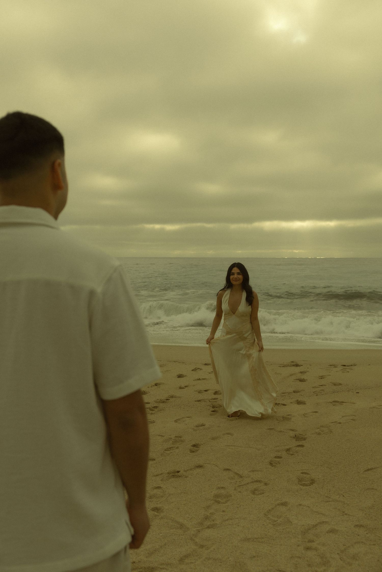 Couple in white attire on a sandy beach, woman in flowing dress walks toward man, ocean waves and cloudy sky behind.
