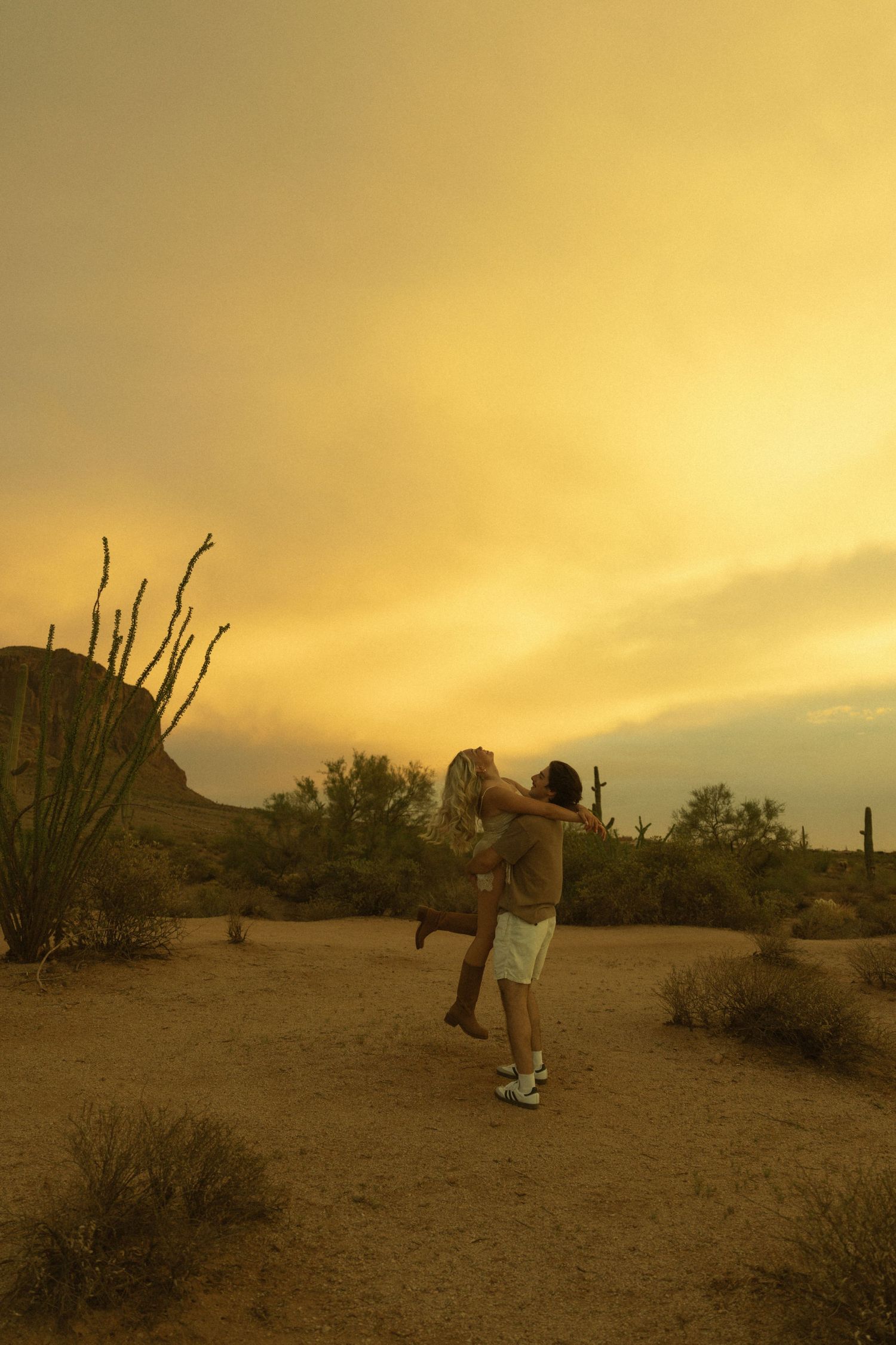 Couple embracing and spinning in a desert landscape at golden sunset with cacti and dramatic sky.